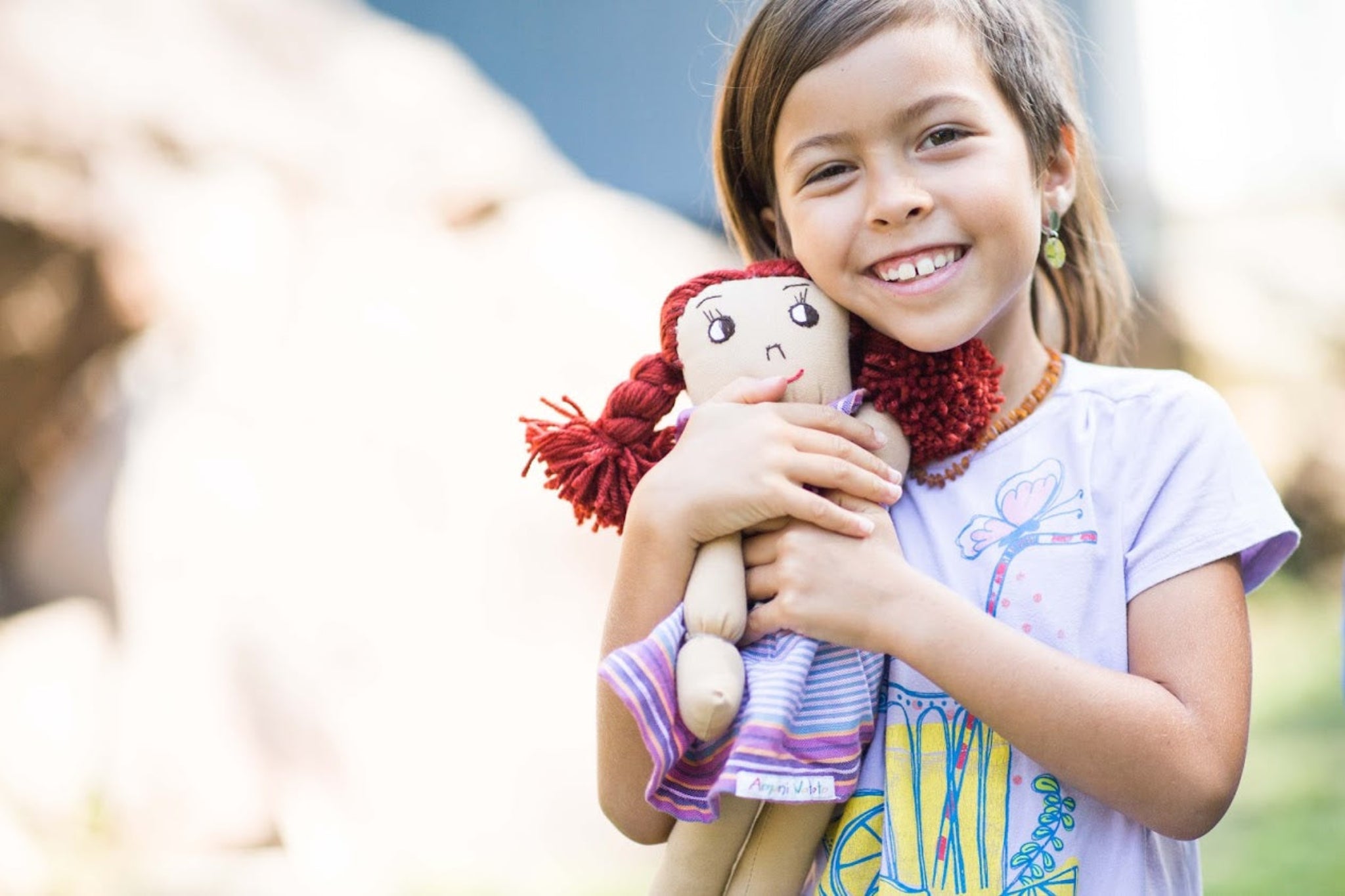 Young girl holding a doll with red hair, smiling outdoors.