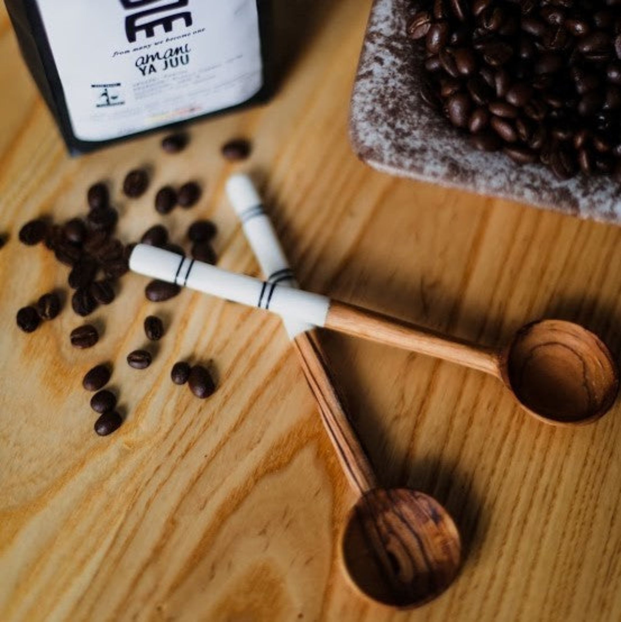 A wooden spoon with a bone handle, displayed on a wooden surface with coffee beans scattered around it. A bag of coffee and a cake are also visible in the background.