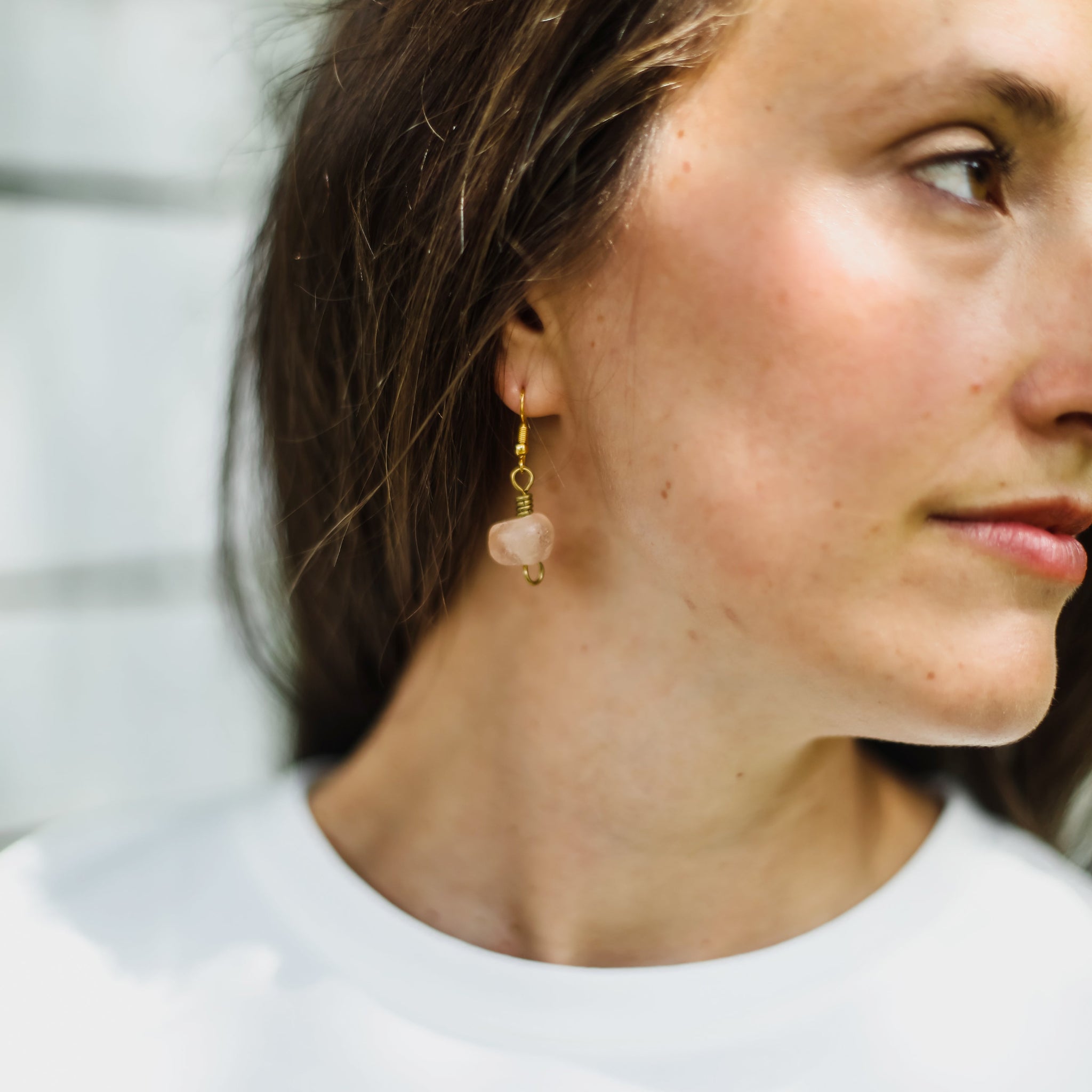 A close-up side profile of a woman wearing a delicate gold dangle earring with a small, polished pink stone charm.