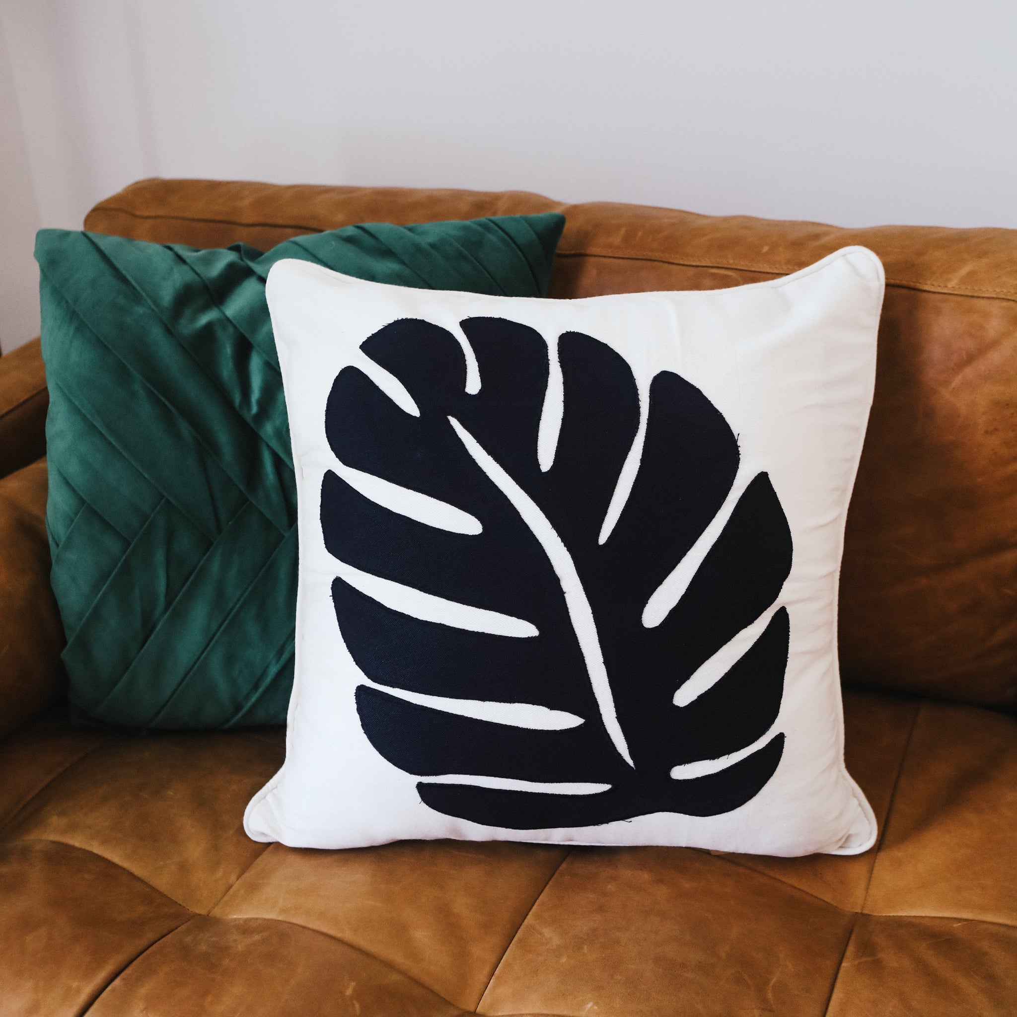 A decorative pillow with a black monstera leaf print on a white background, displayed on a brown couch with a green cushion.