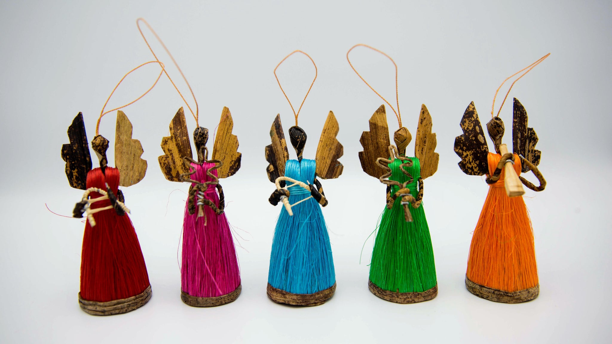 Five small handmade angel ornaments standing in a row on a light gray background. Each angel has a different colored dress: red, magenta, blue, green, and orange, with brown and gold wings and a hanging string.