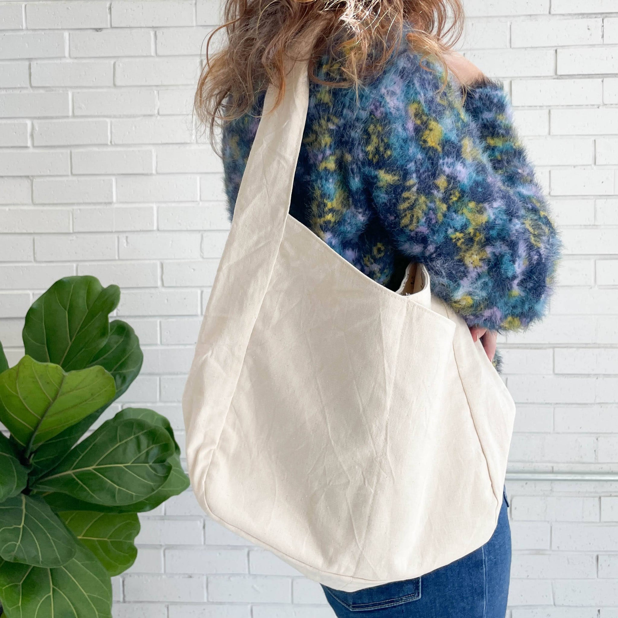 Person holding a beige tote bag against a white brick wall with a plant in the foreground.