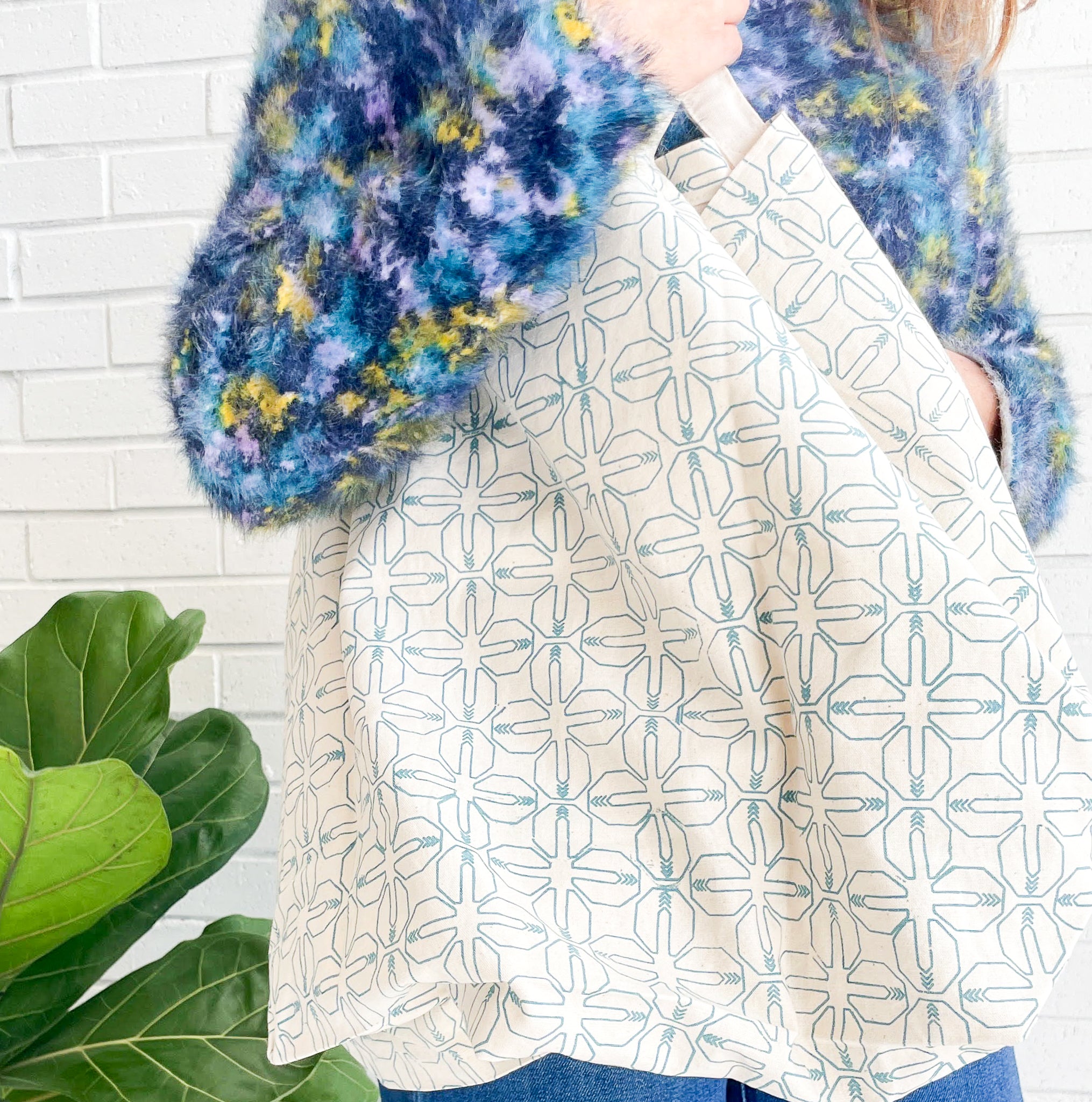 Person wearing a blue and yellow patterned sweater holding a white tote bag with a geometric design, standing against a white brick wall with a plant in the foreground.
