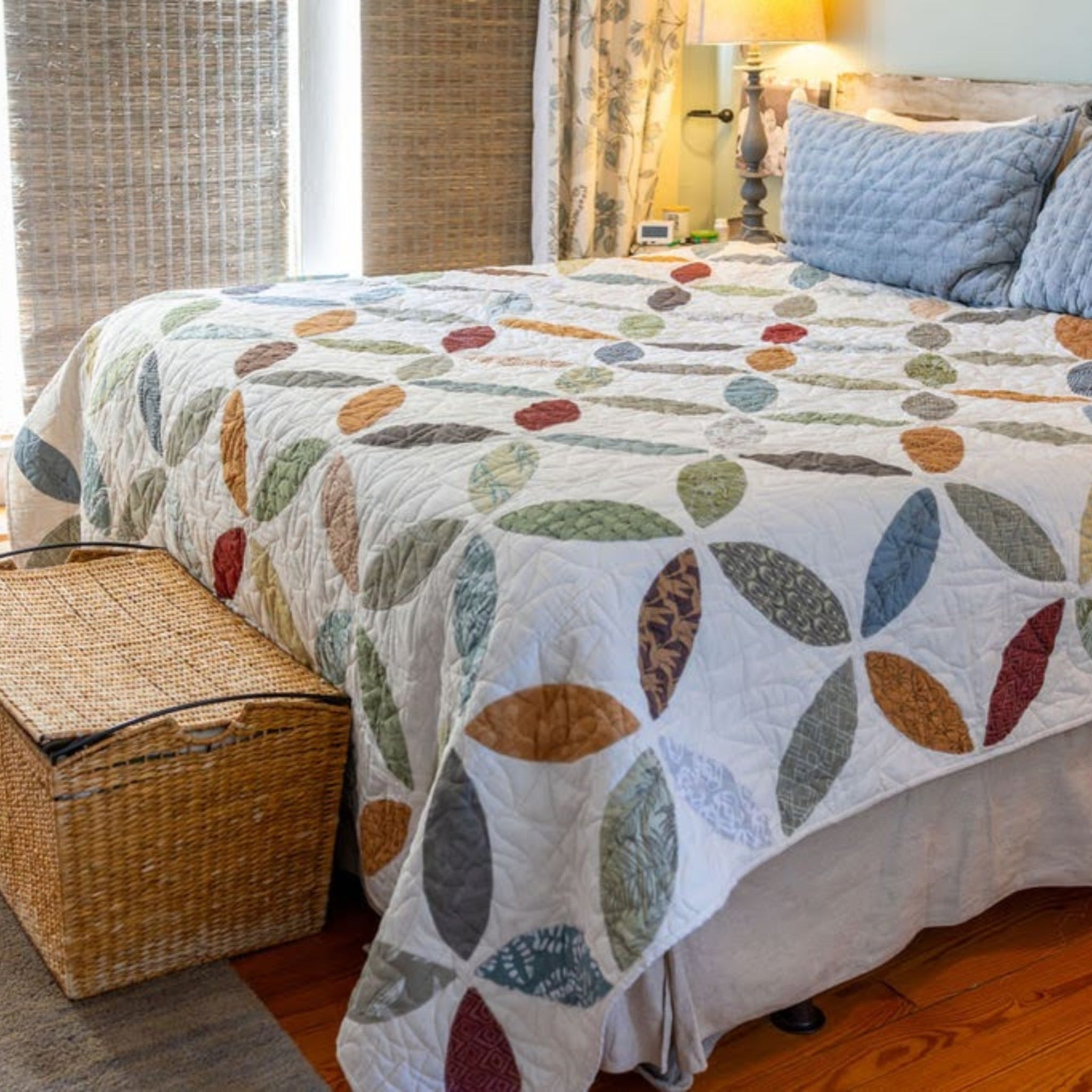 A colorful quilted bedspread with a repeating petal pattern in muted blue, green, brown, and rust tones, styled on a bed in a bedroom with two blue pillows, a wicker bench, and wooden floor.