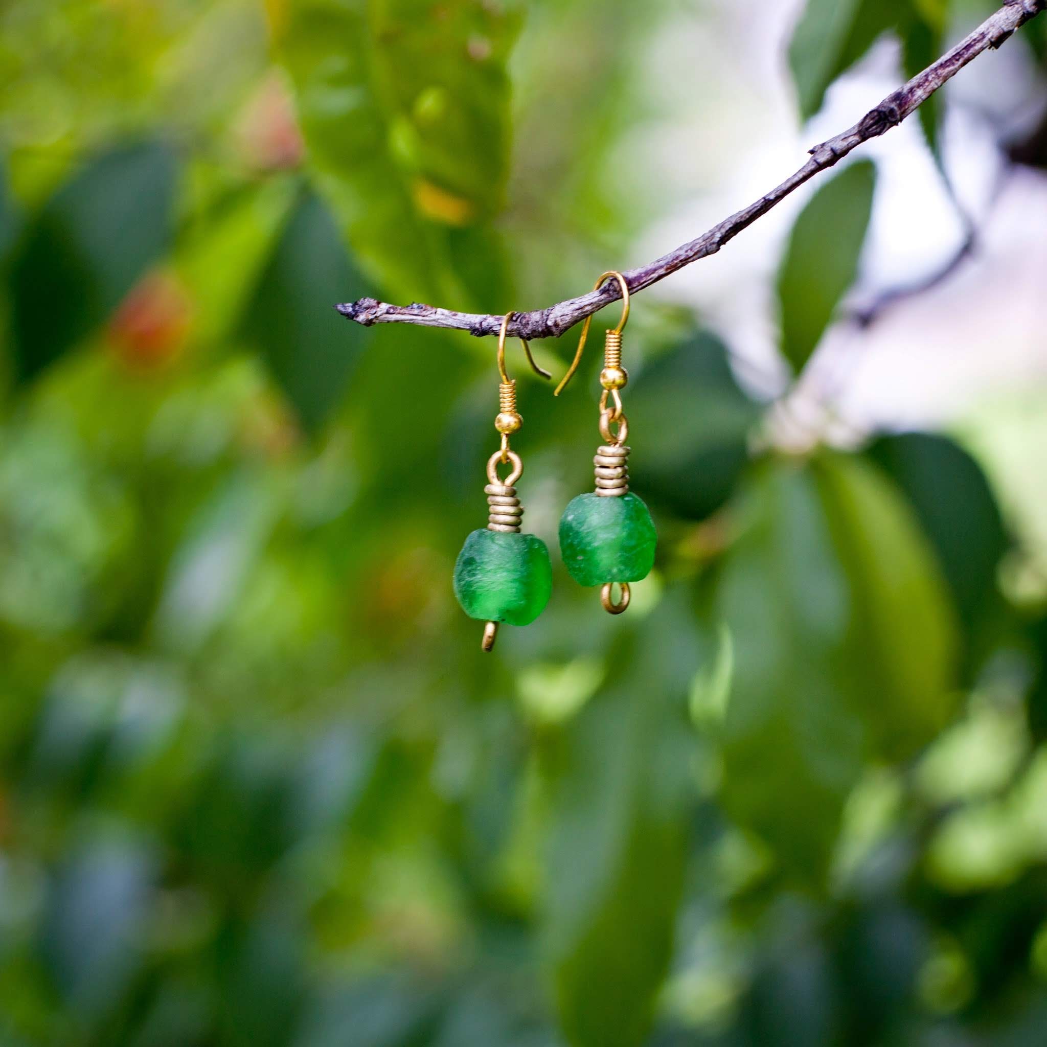 A pair of handmade drop earrings with translucent green beads and gold wire-wrapped hooks, hanging from a thin tree branch against a soft-focus background of green leaves.