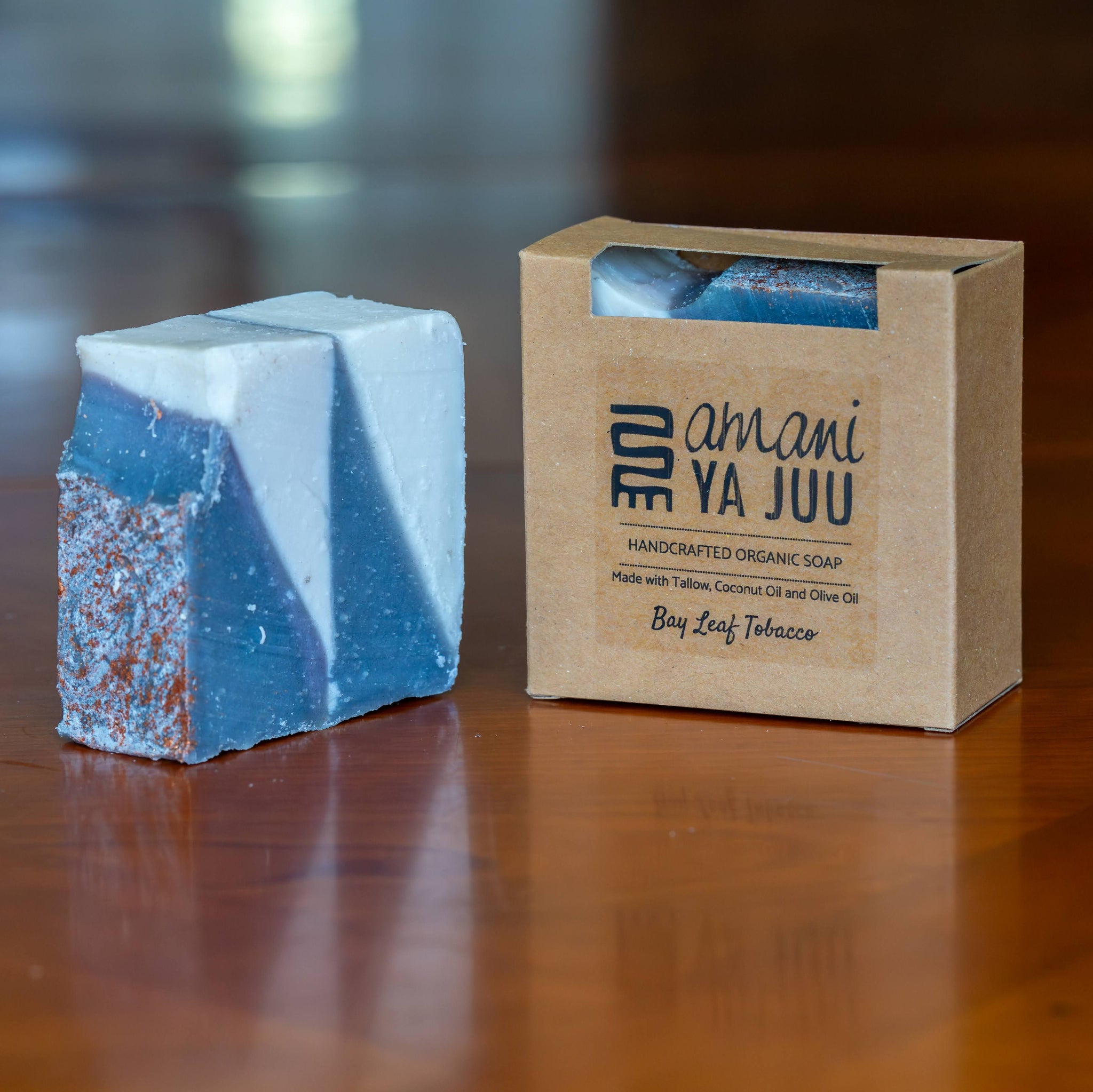 A handmade soap bar with a blue and white marbled pattern and coppery speckles, standing next to its brown kraft paper packaging box labeled 'amani YA JUU' on a wooden table.