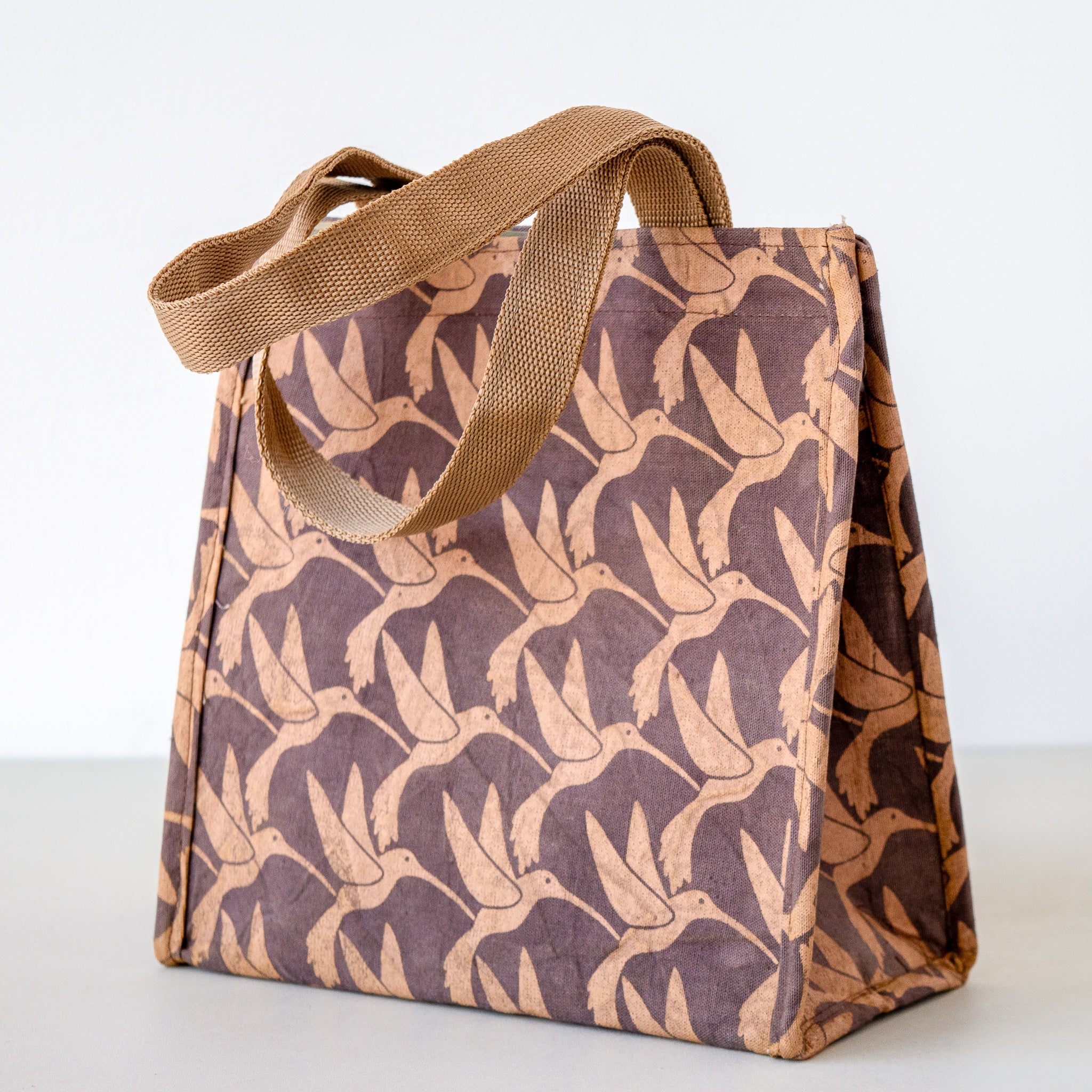 A square canvas lunch bag with a brown and tan humming bird pattern, featuring thick tan shoulder straps, photographed on a light gray background.