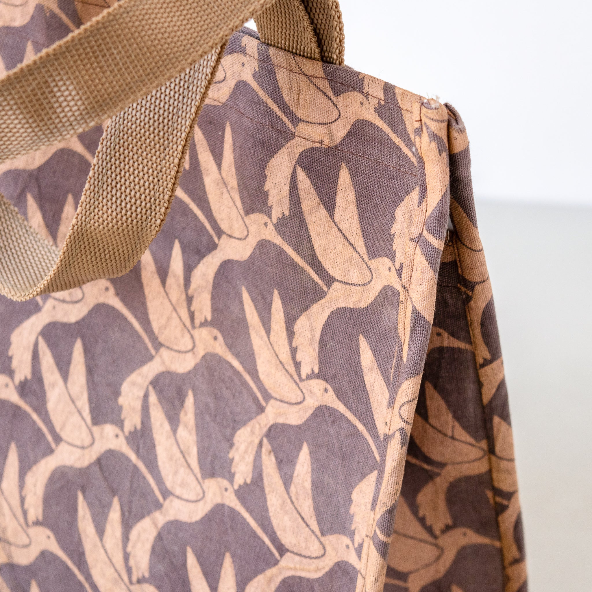 Close-up of a fabric tote bag with a repeating humming bird print in tan and dark brown, with a woven tan strap visible at the top left.