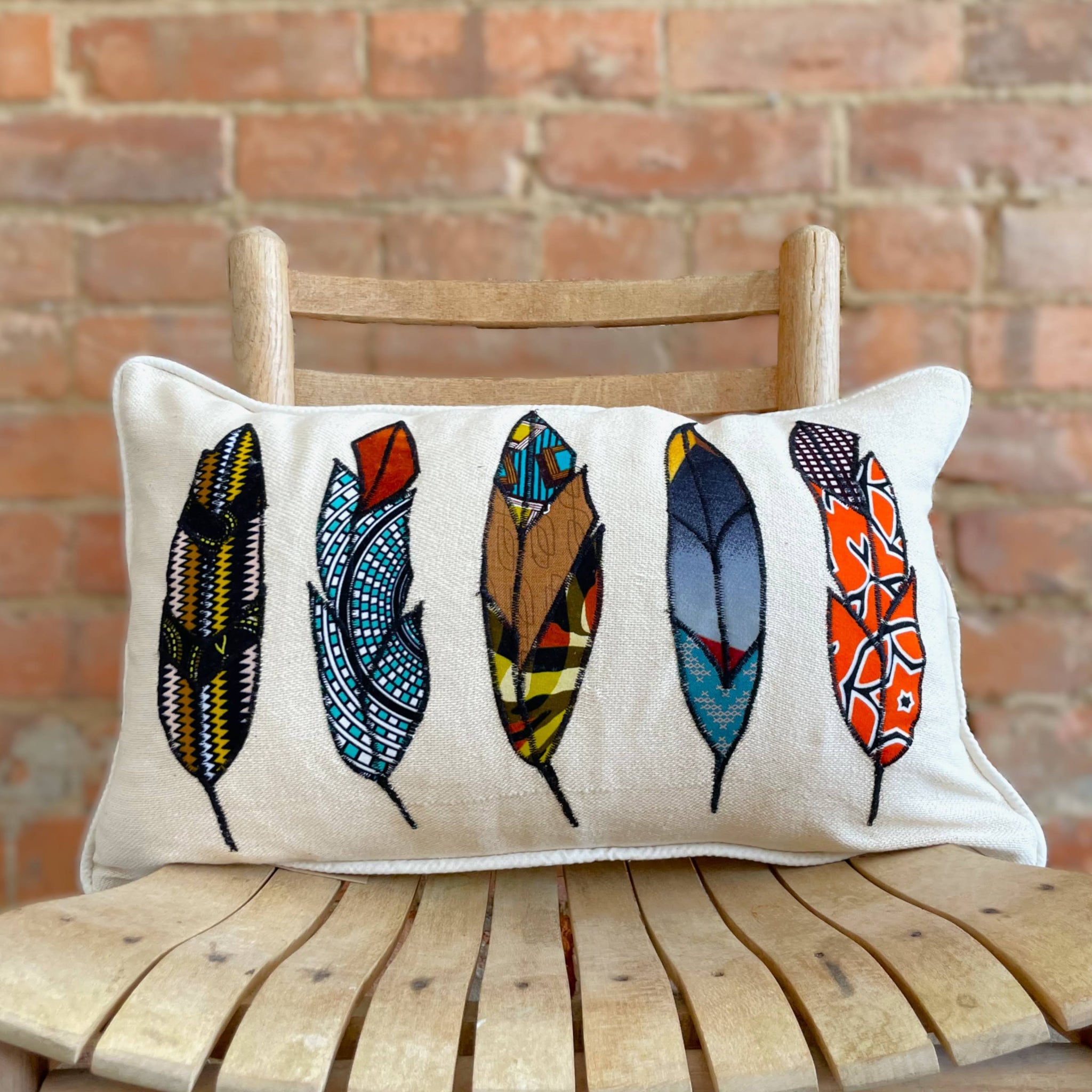 Decorative pillow with colorful feather designs on a wooden chair against a brick wall.