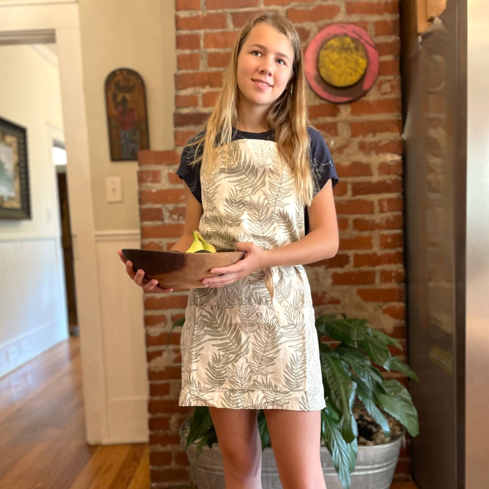 Person wearing a fern-patterned apron holding a bowl in a home setting with a brick wall.