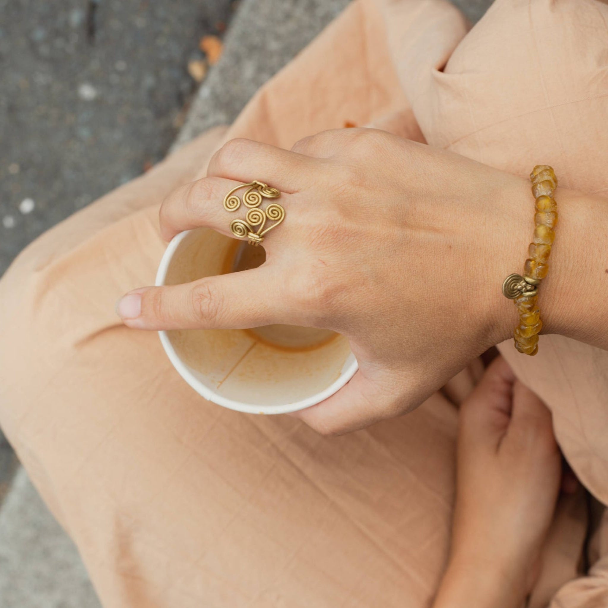 Person holding a cup with an adjustable brass ring and bottle bead bracelet on a blurred background
