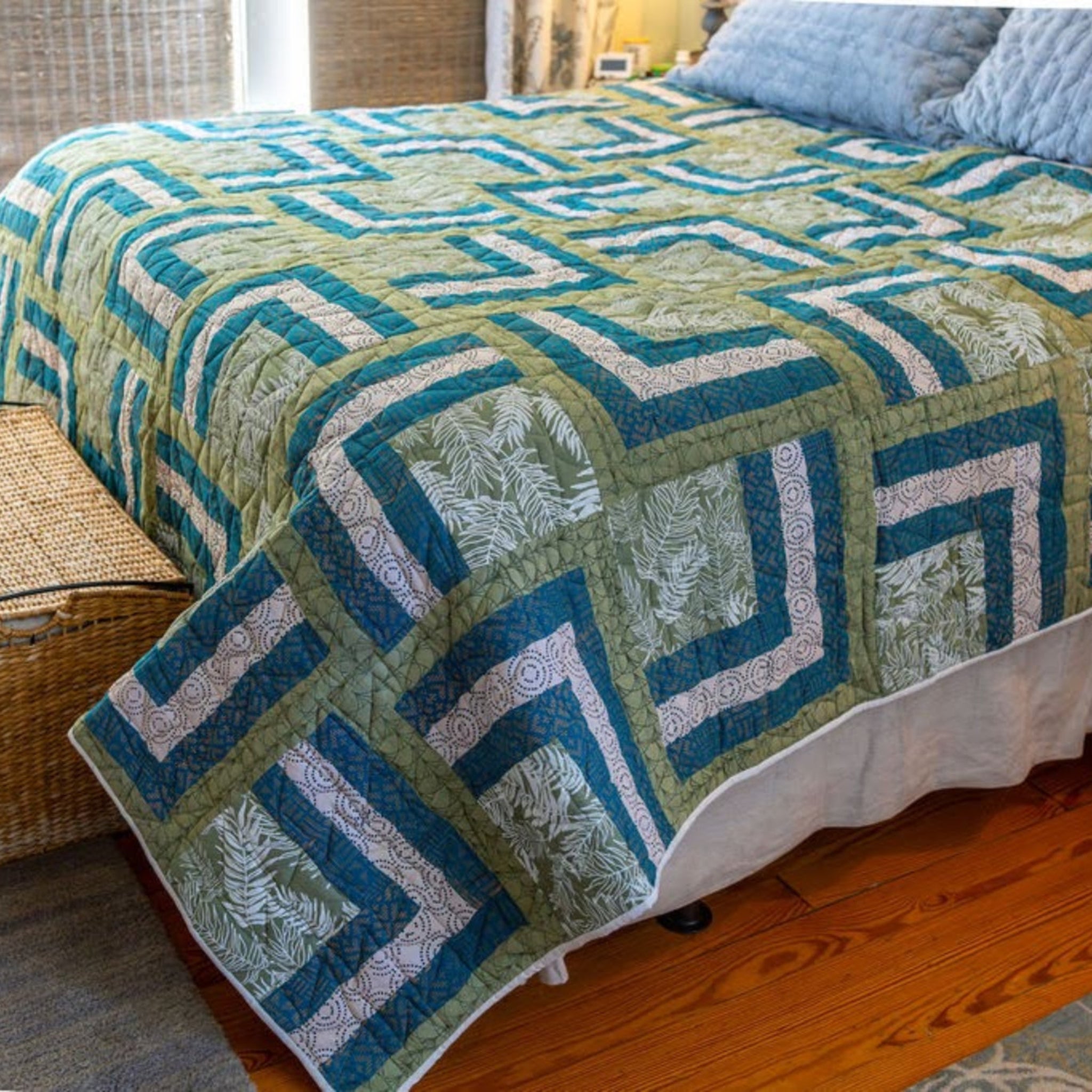 A quilted patchwork bedspread with a geometric and botanical pattern in teal, sage green, and cream, styled on a bed in a bedroom setting.