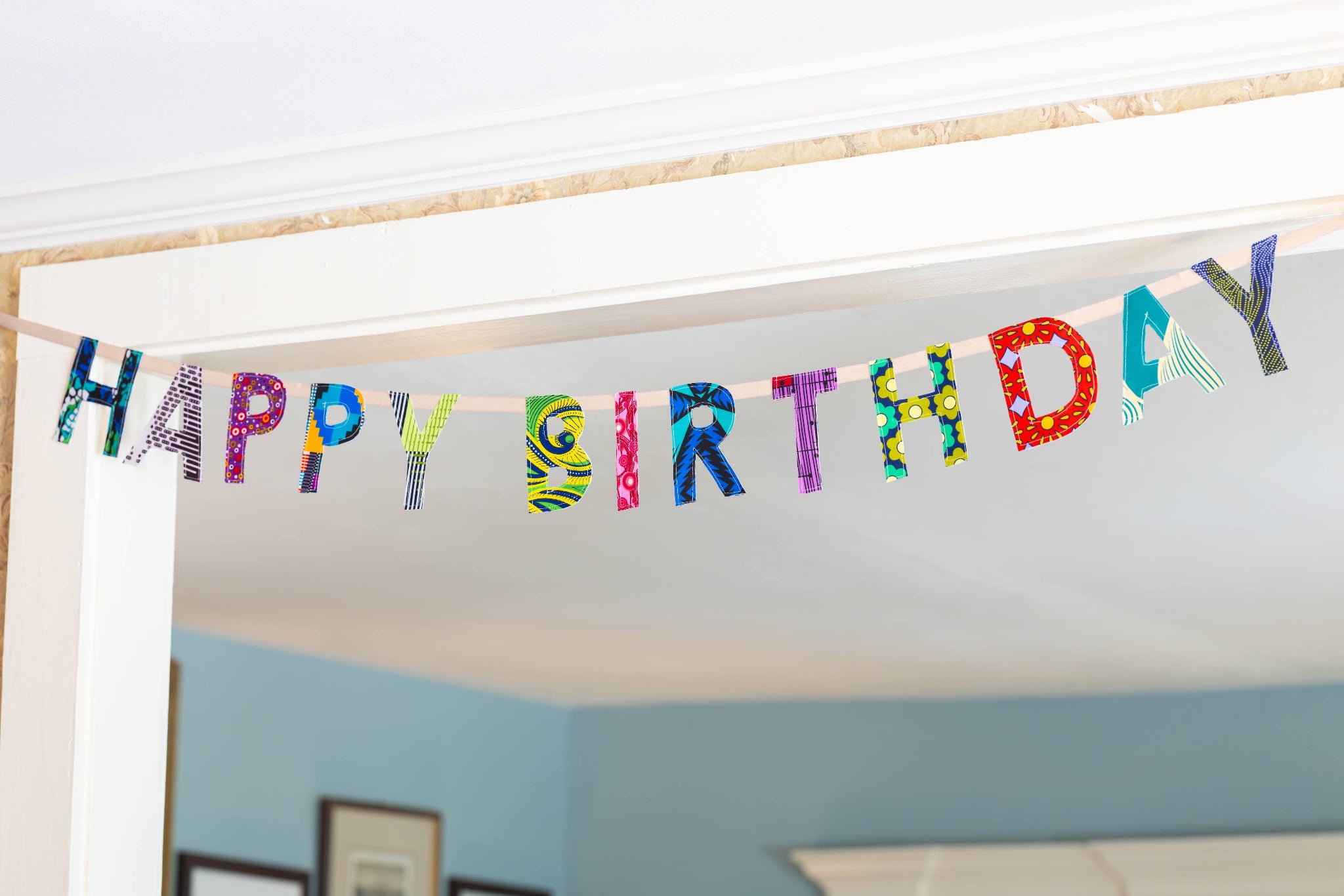 A colorful 'HAPPY BIRTHDAY' banner made of patterned paper letters hanging across a doorway in a home interior with light blue walls.