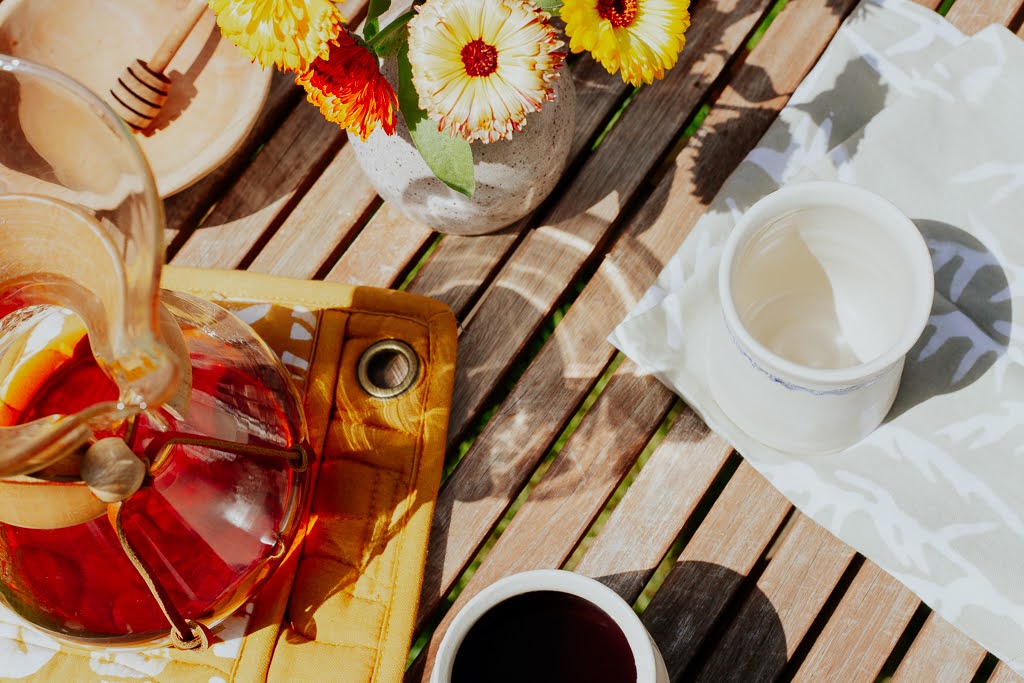 Outdoor setting with a glass pitcher of iced tea, a white mug, and colorful flowers on a wooden table.