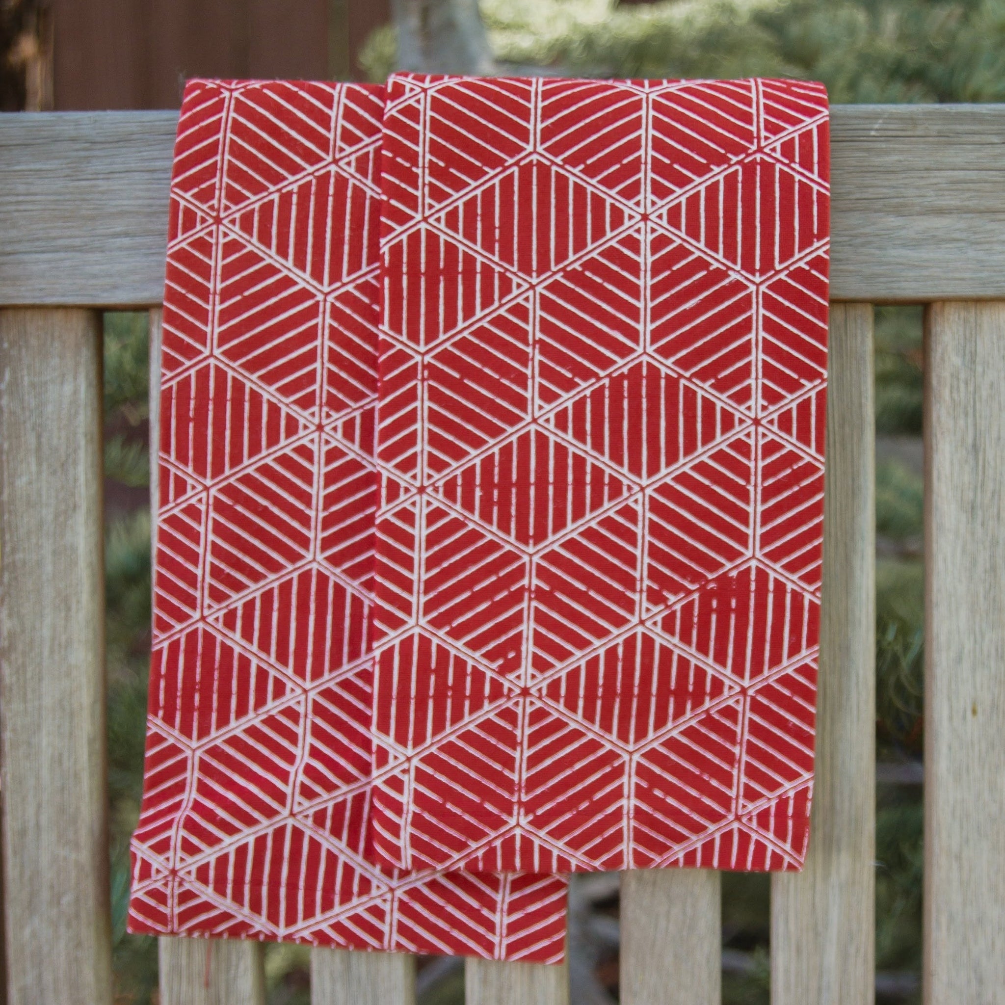 Red geometric patterned tea towels hanging on a wooden fence with blurred greenery in the background