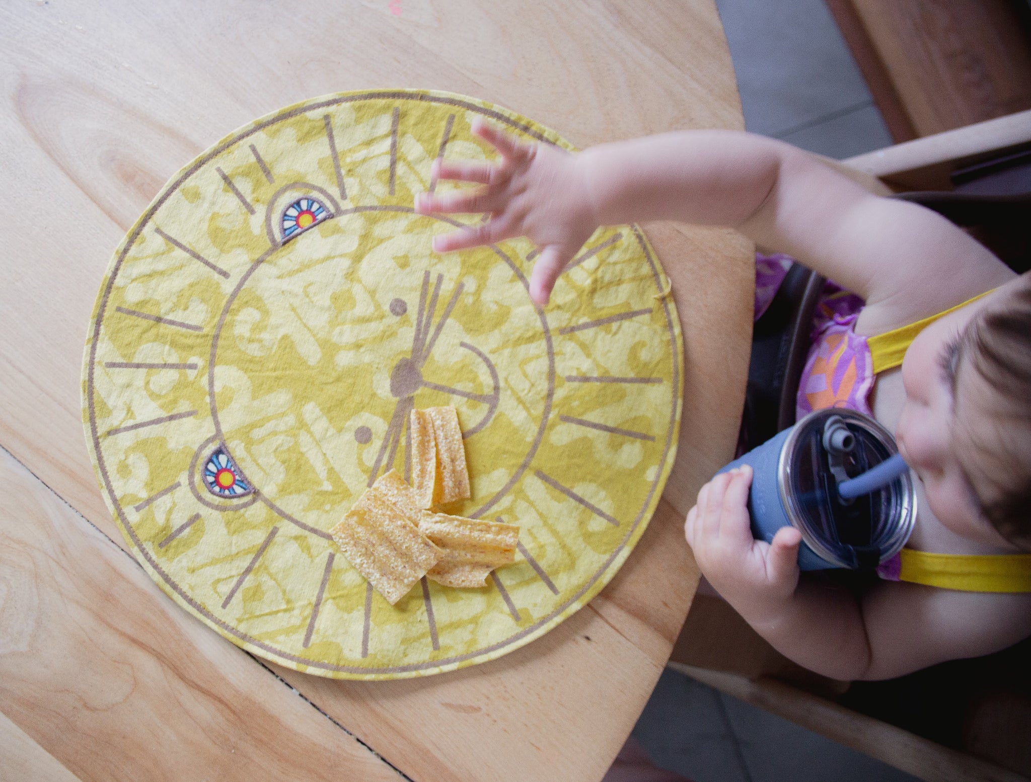 Child's hand reaching towards a round yellow placemat with a lion face design on a wooden table.