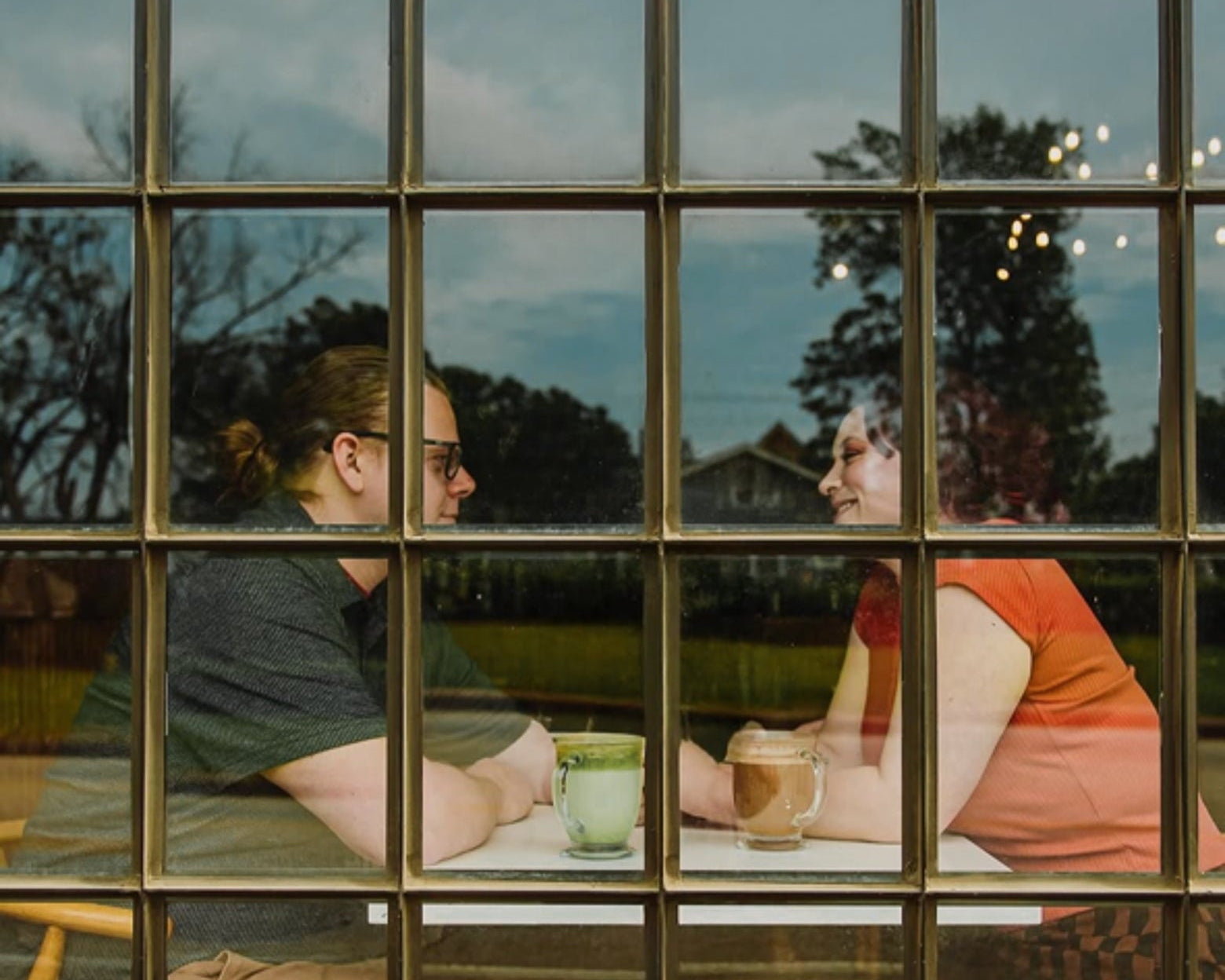 Two people sitting at a table with drinks, viewed through a window.