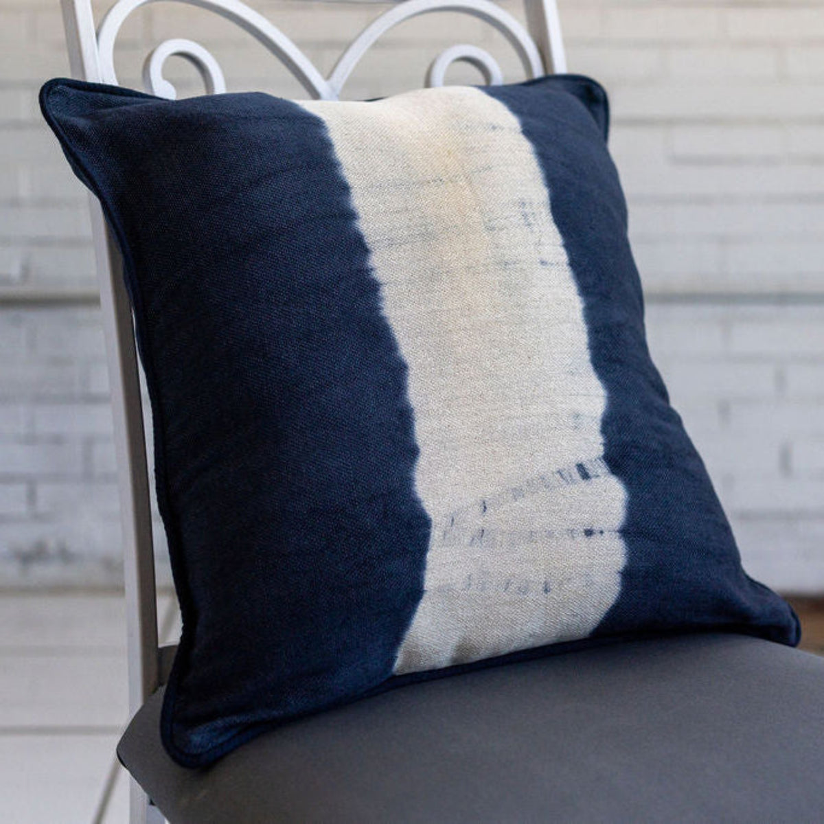 A square throw pillow with a navy blue and off-white vertical tie-dye style stripe pattern, resting on a gray cushioned chair in front of a white brick wall.
