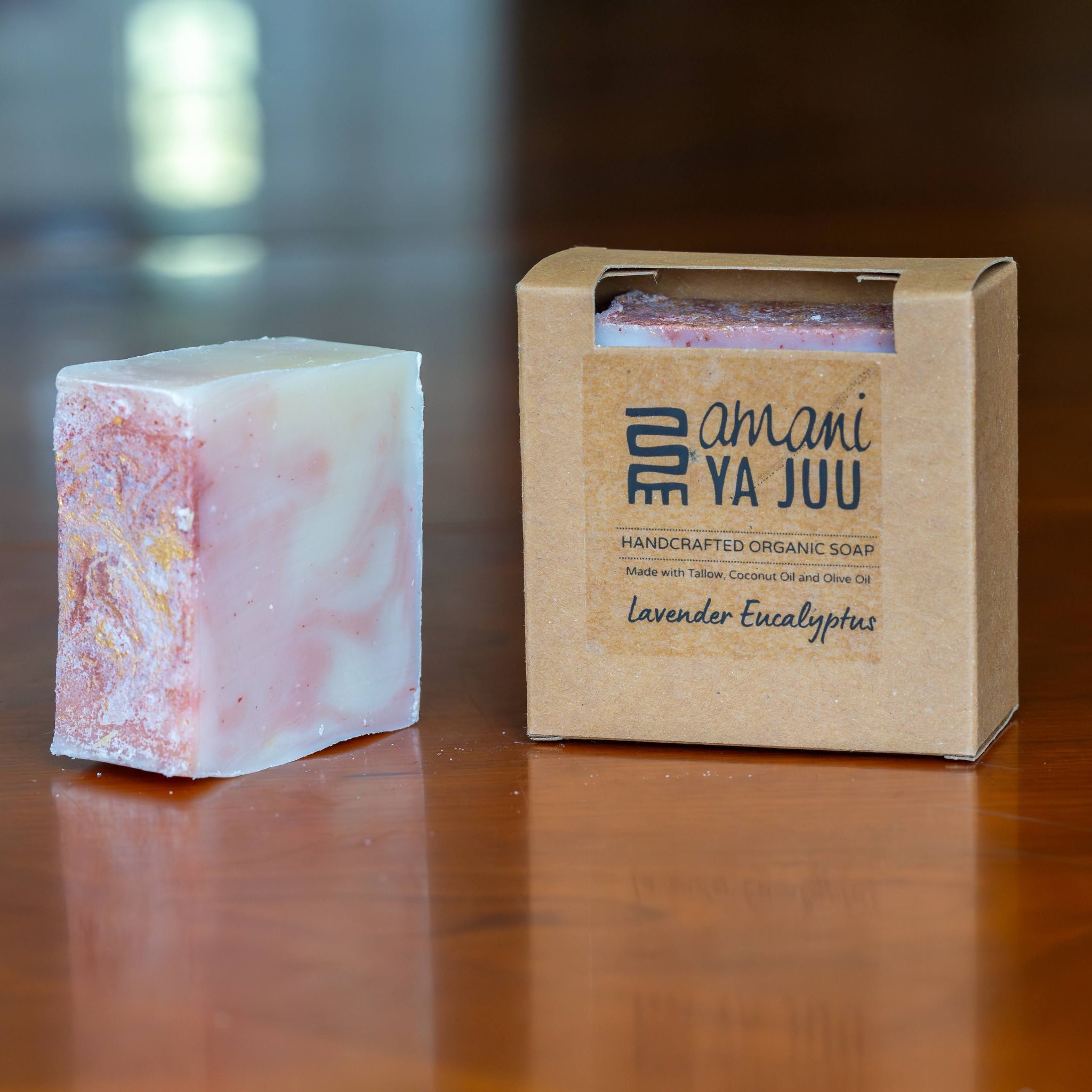 A handmade soap bar with pink and white marbling sits next to its kraft paper packaging box on a polished wooden table. The box is branded 'amani YA JUU' and labeled 'Lavender Eucalyptus'.