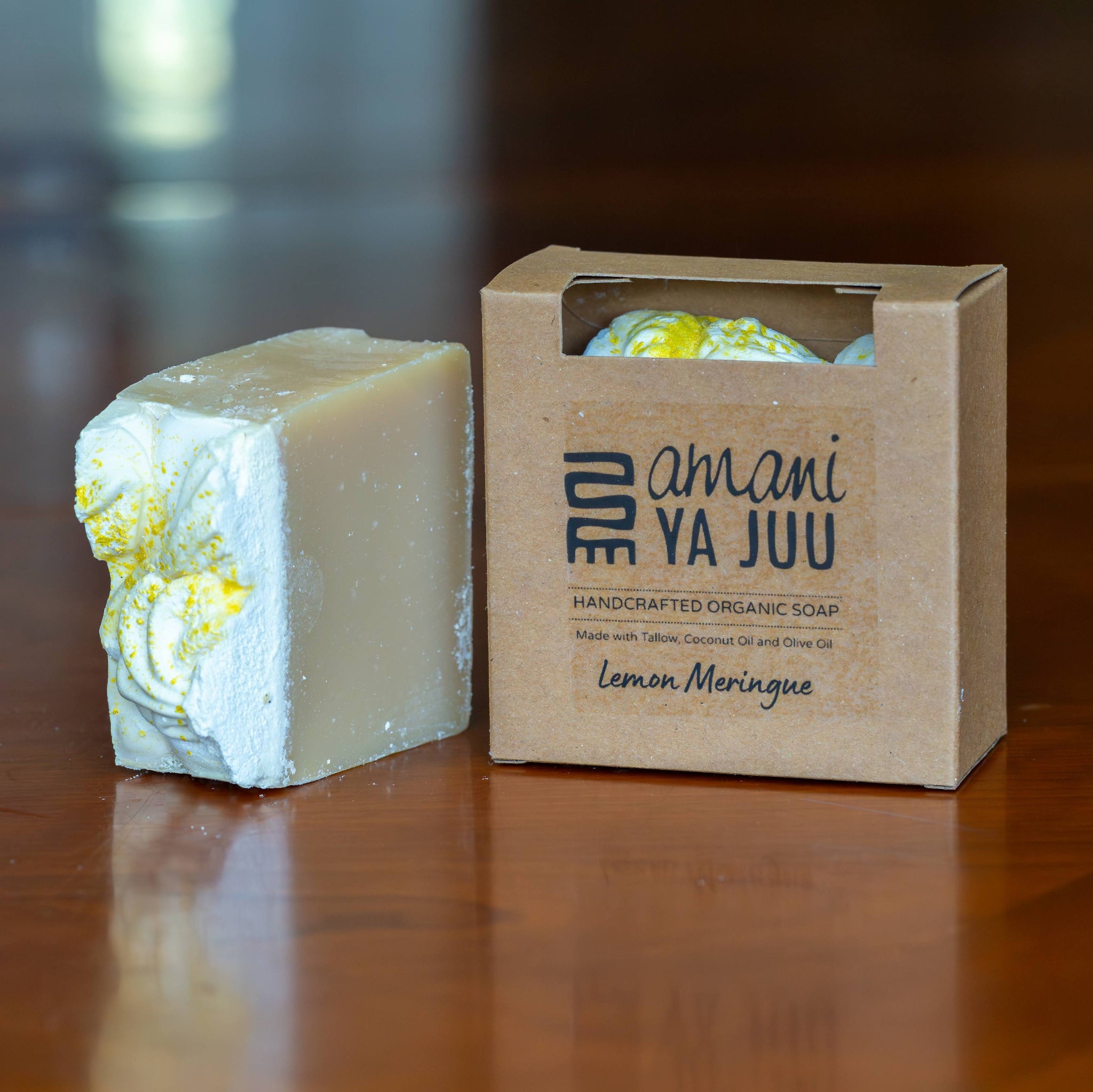 A handmade organic soap bar with yellow lemon swirls next to its kraft paper packaging box labeled 'amani YA JUU' on a wooden table.