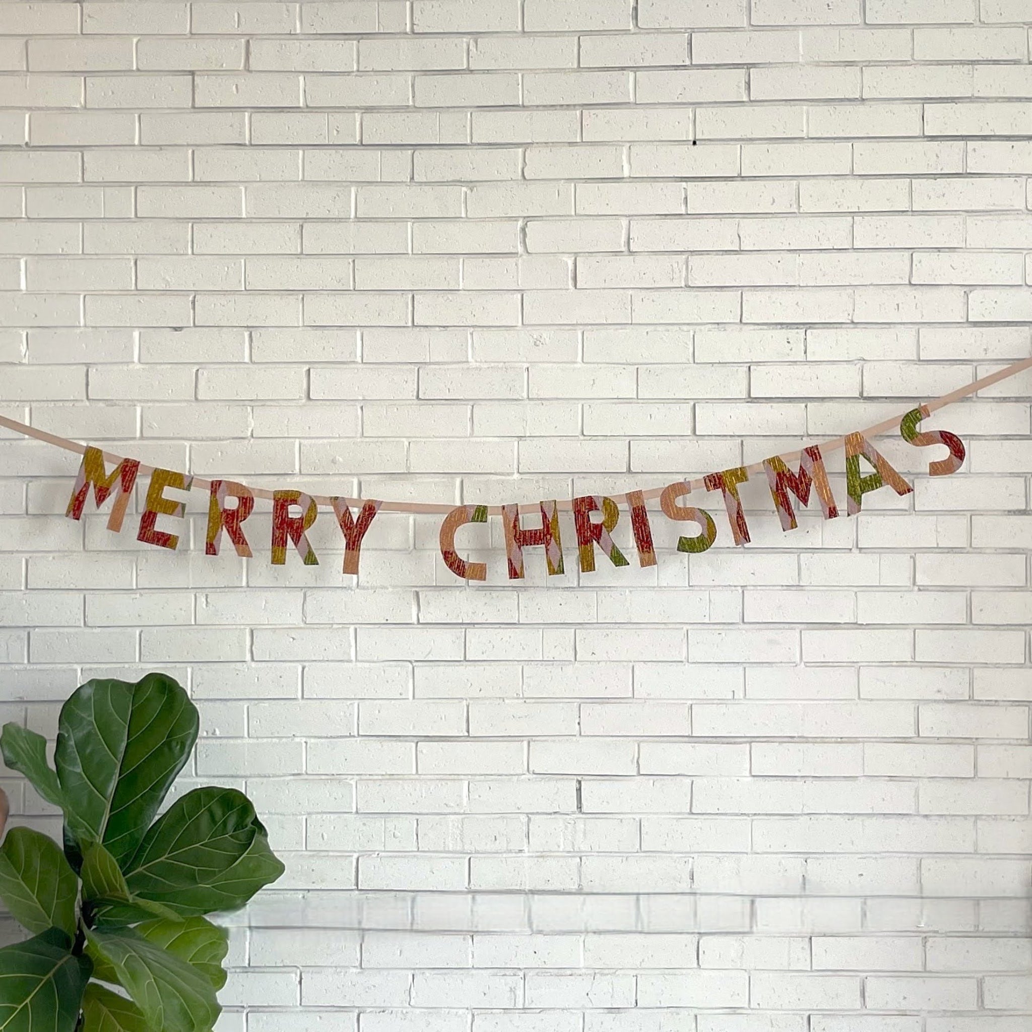 Merry Christmas banner on a white brick wall with a plant in the corner.