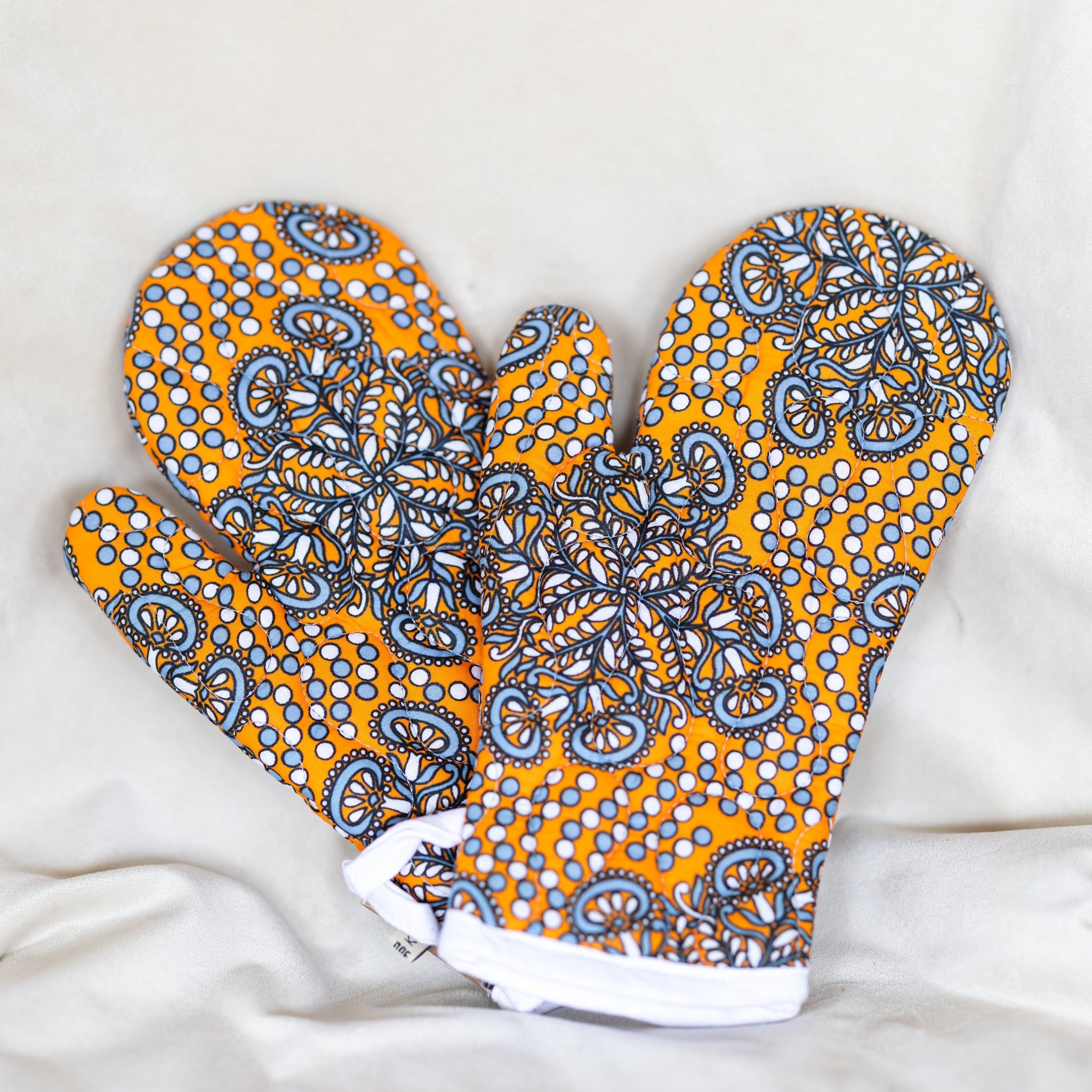 A pair of oven mitts featuring a vibrant yellow-orange base with a detailed blue, gray, and white paisley pattern, laid flat on a white fabric background.