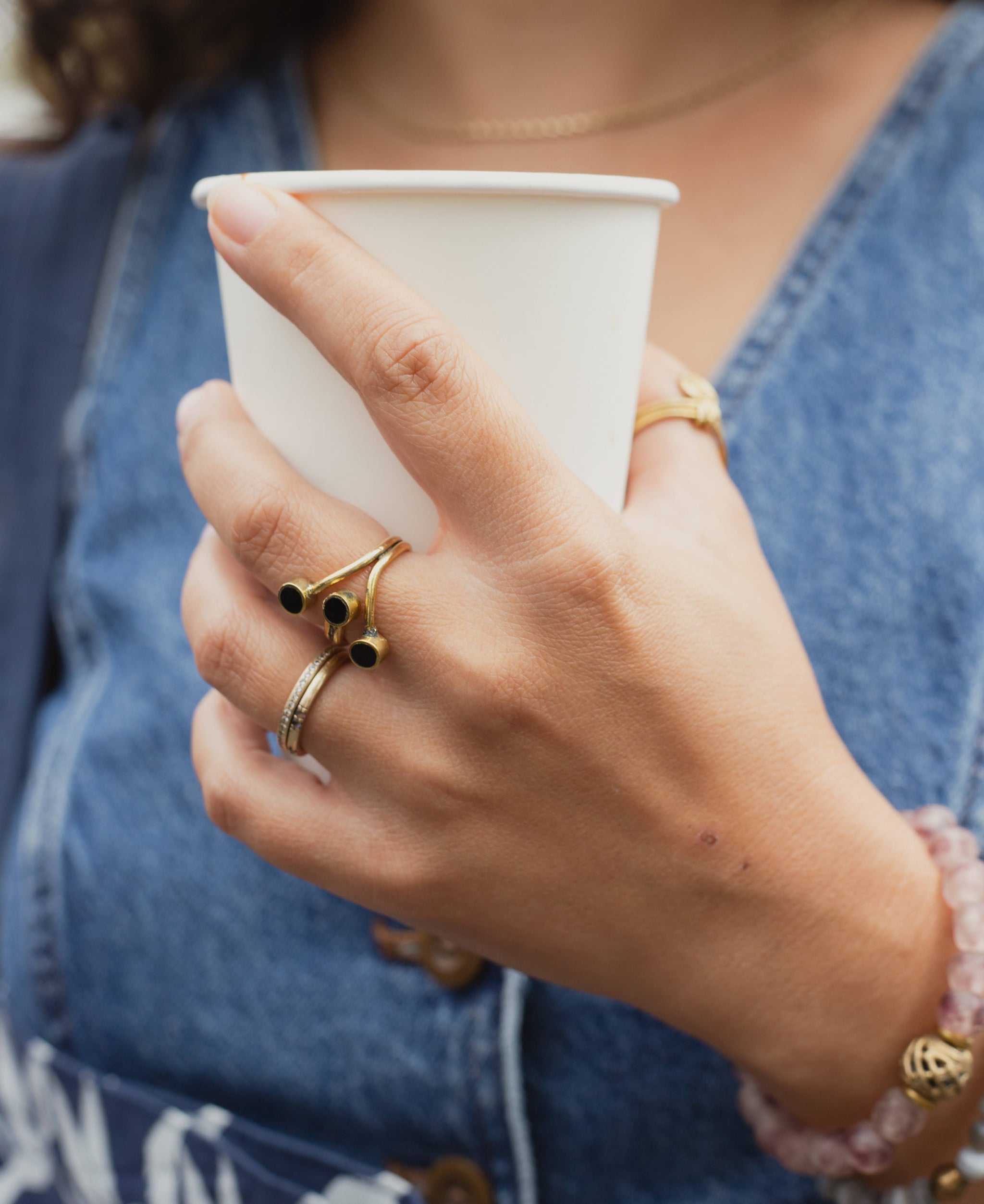 Hand holding a white paper cup with brass and horn ring on finger and bottle bead bracelets on wrist