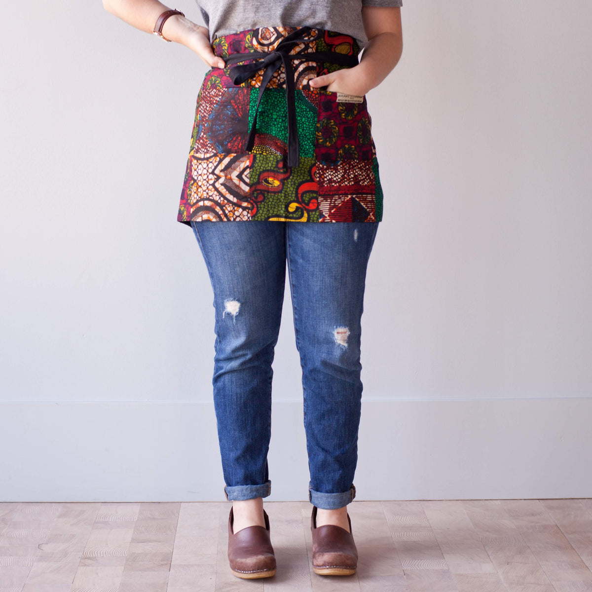 Person wearing jeans, grey shirt, and multicolored patch half apron tied at the waist in front of a light background.