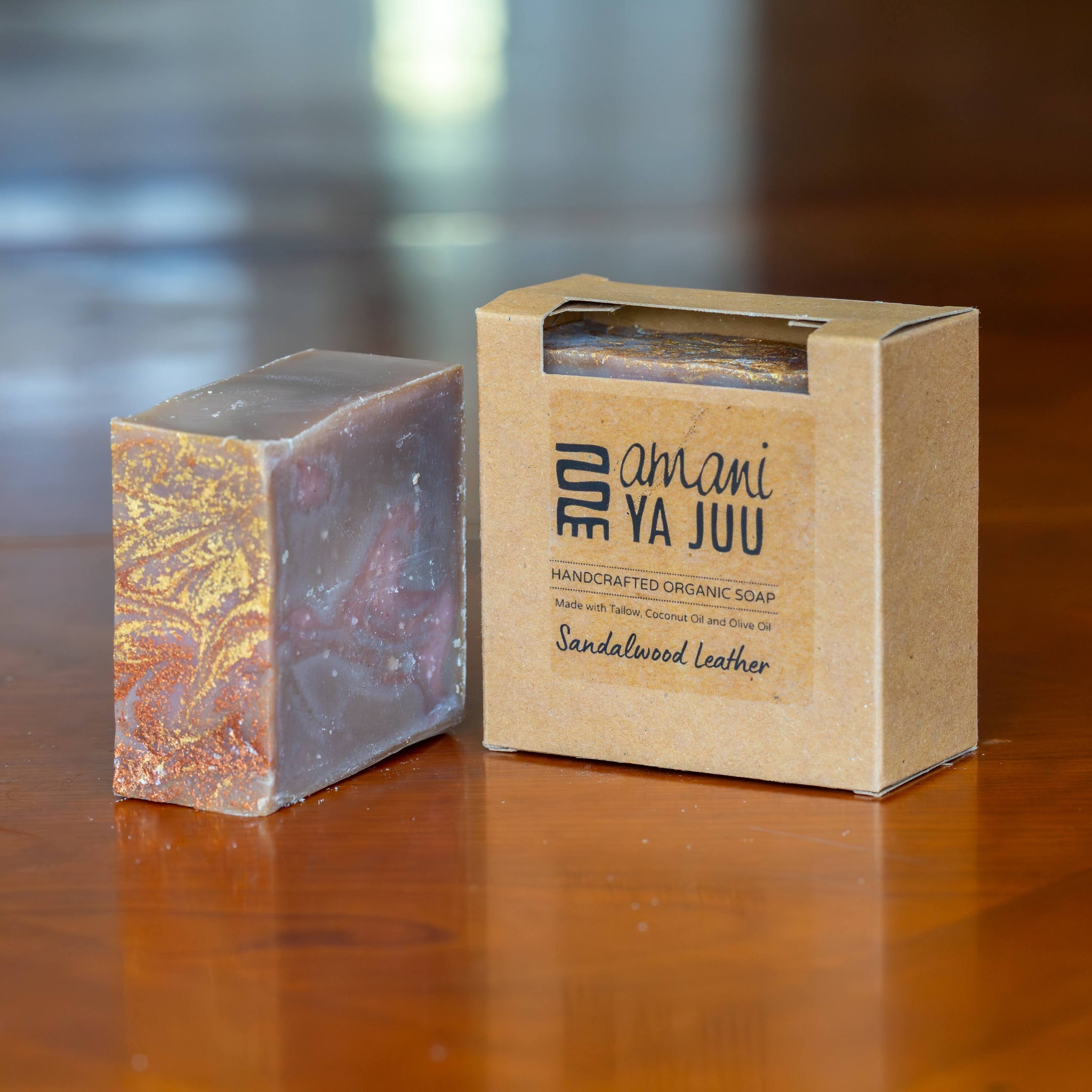 A handmade soap bar with a marbled purple, gold, and orange pattern stands next to its kraft paper packaging box on a polished wooden surface. The box is labeled 'amani YA JUU' and 'Sandalwood Leather'.