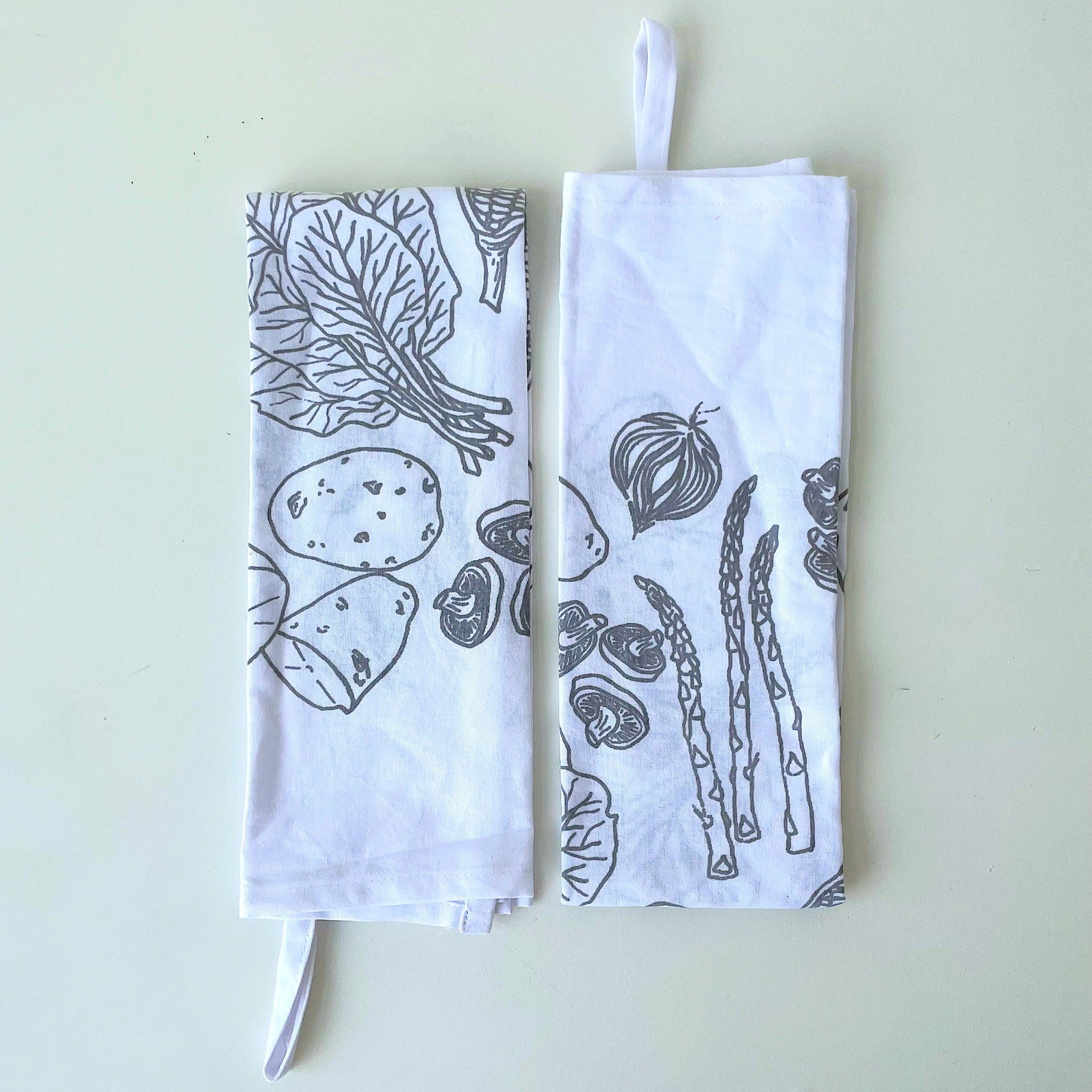 Two white towels with sage grey vegetable illustrations on a light background