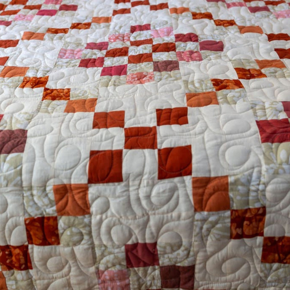 A close-up of a quilted patchwork blanket or quilt featuring alternating white blocks with swirling quilting and orange, red, pink, and rust patterned squares.