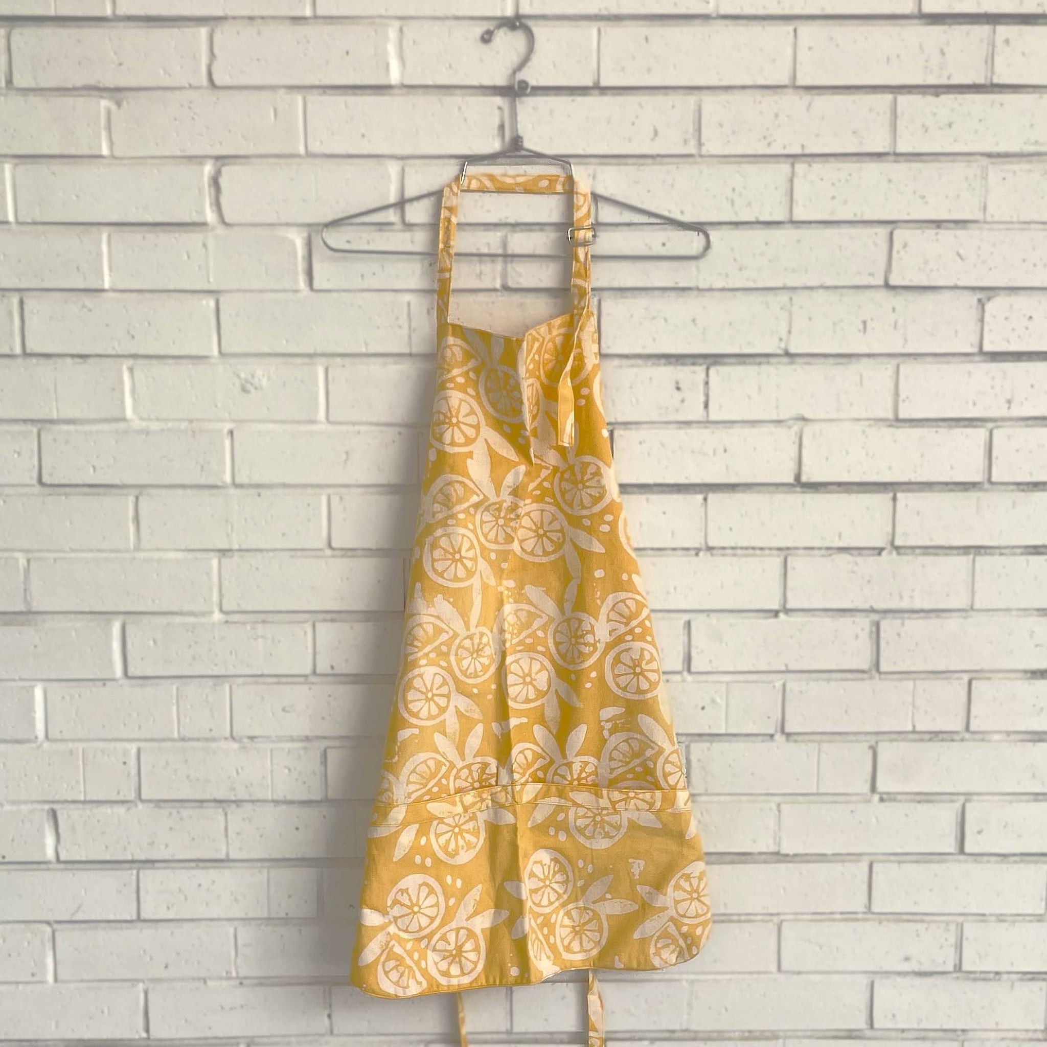Yellow apron with fruit pattern on a hanger against a light brick wall.