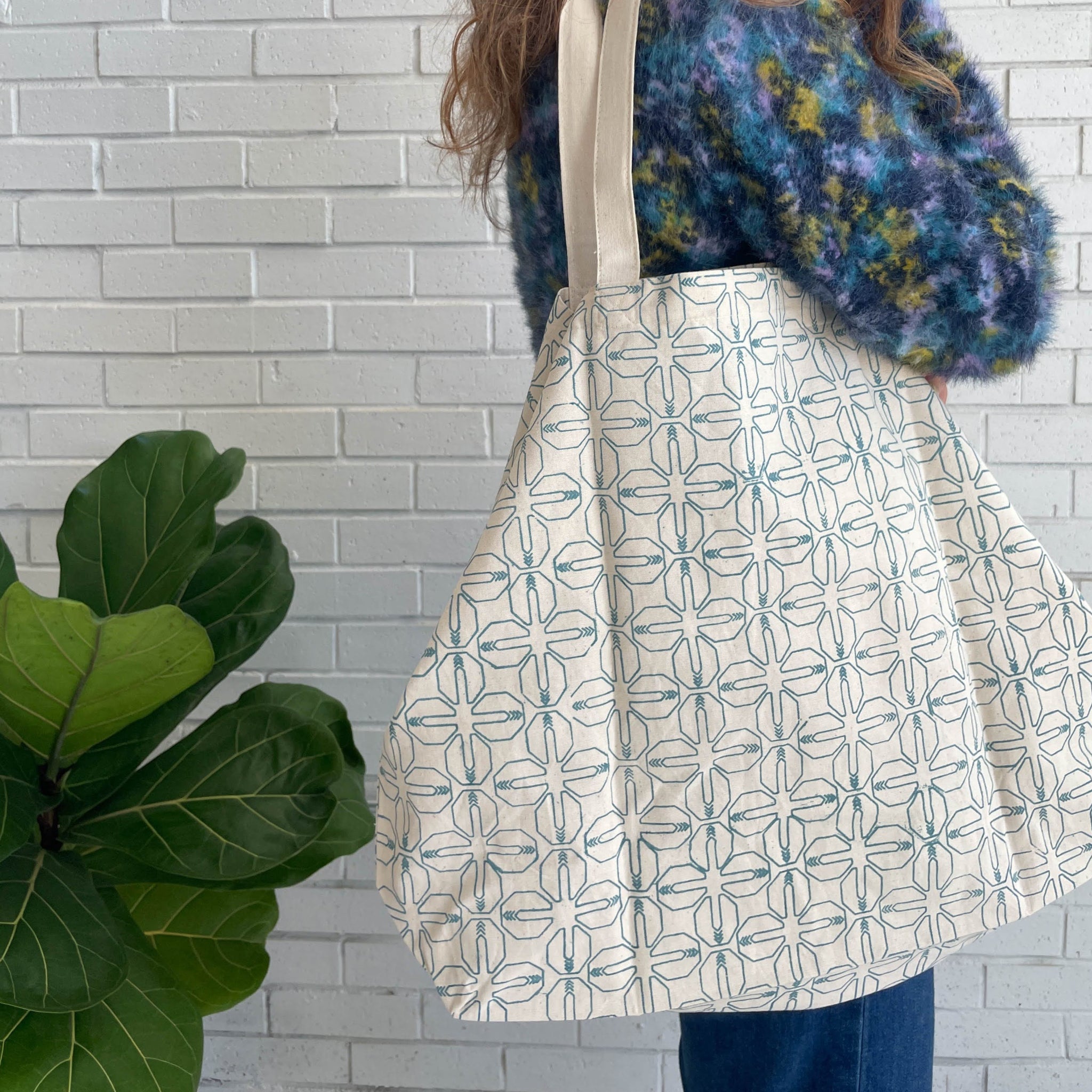 Person holding a patterned tote bag against a white brick wall with a plant in the foreground.