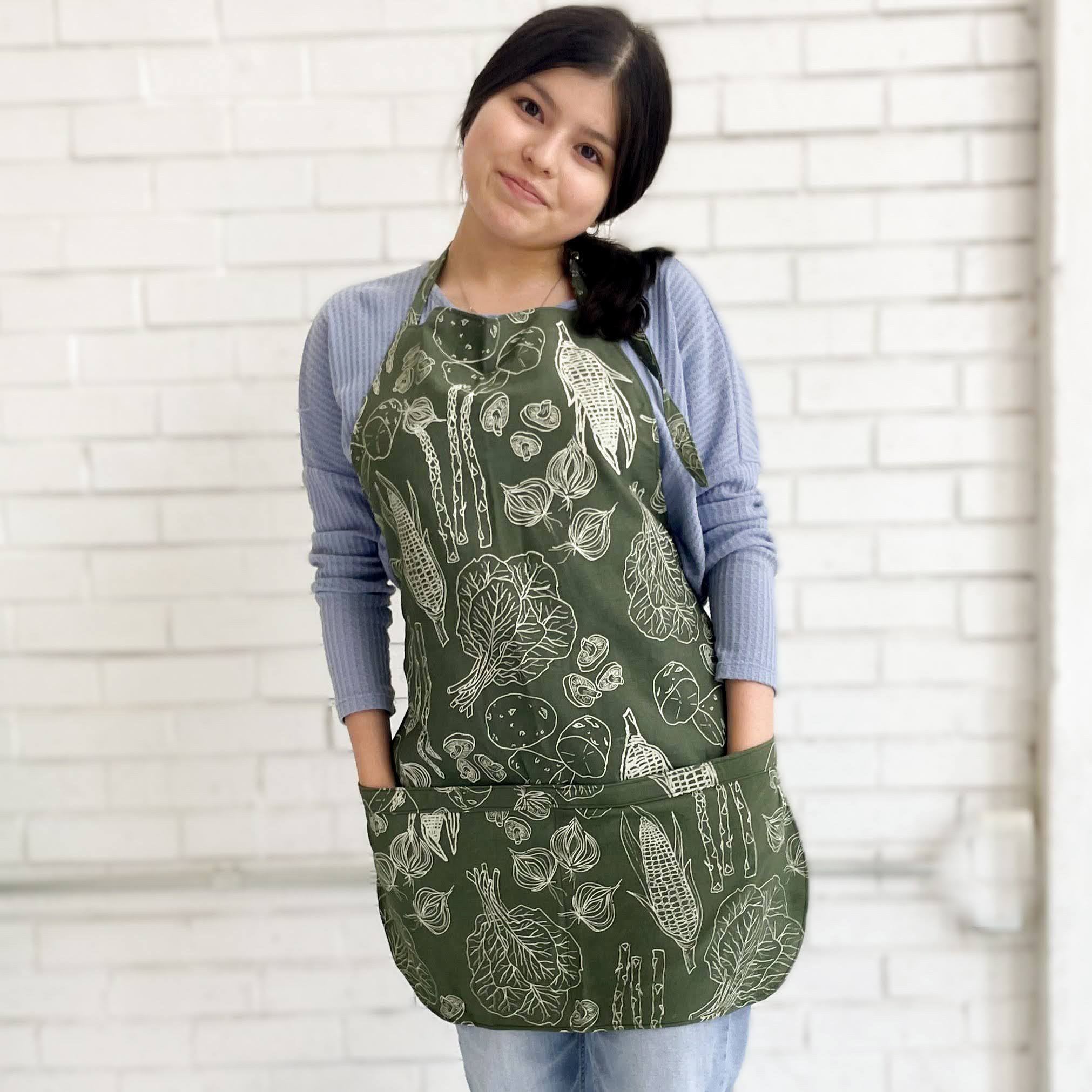 Dark-haired young woman wearing olive green vegetable patterned apron.