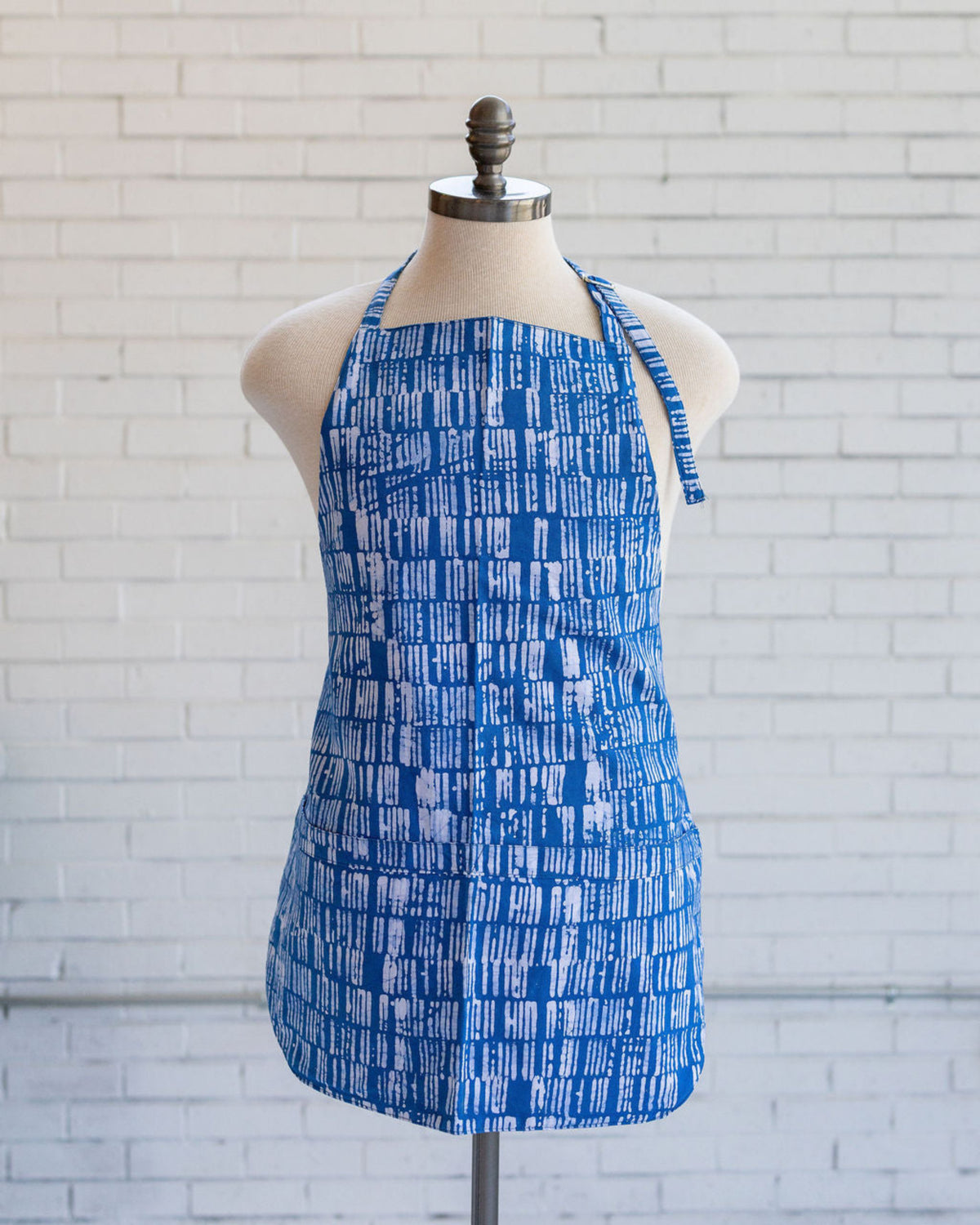 Mannequin displaying a blue and white batik printed striped apron in front of white brick wall