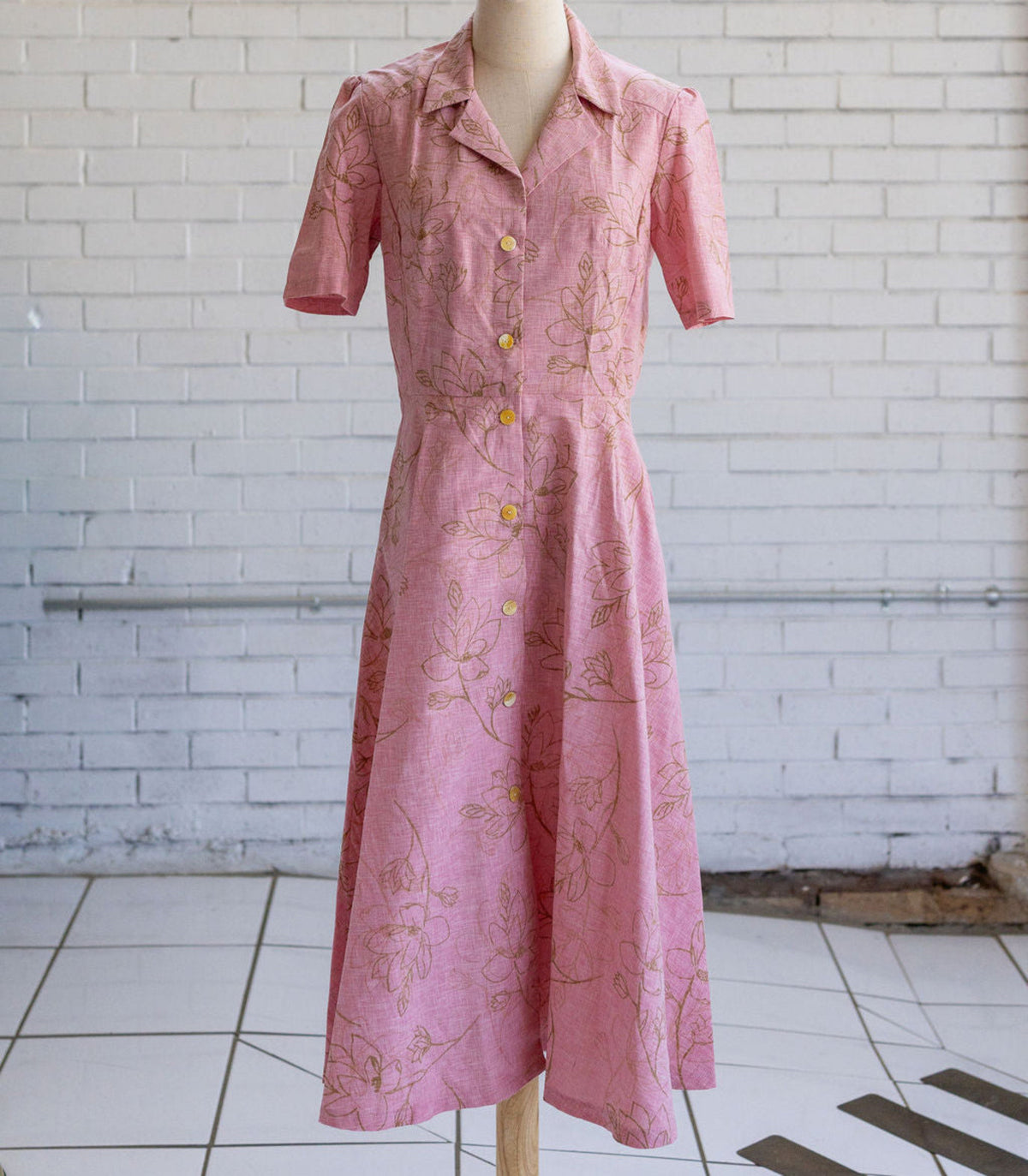 Mannequin wearing a pink button up dress with tan floral pattern in front of white brick wall.