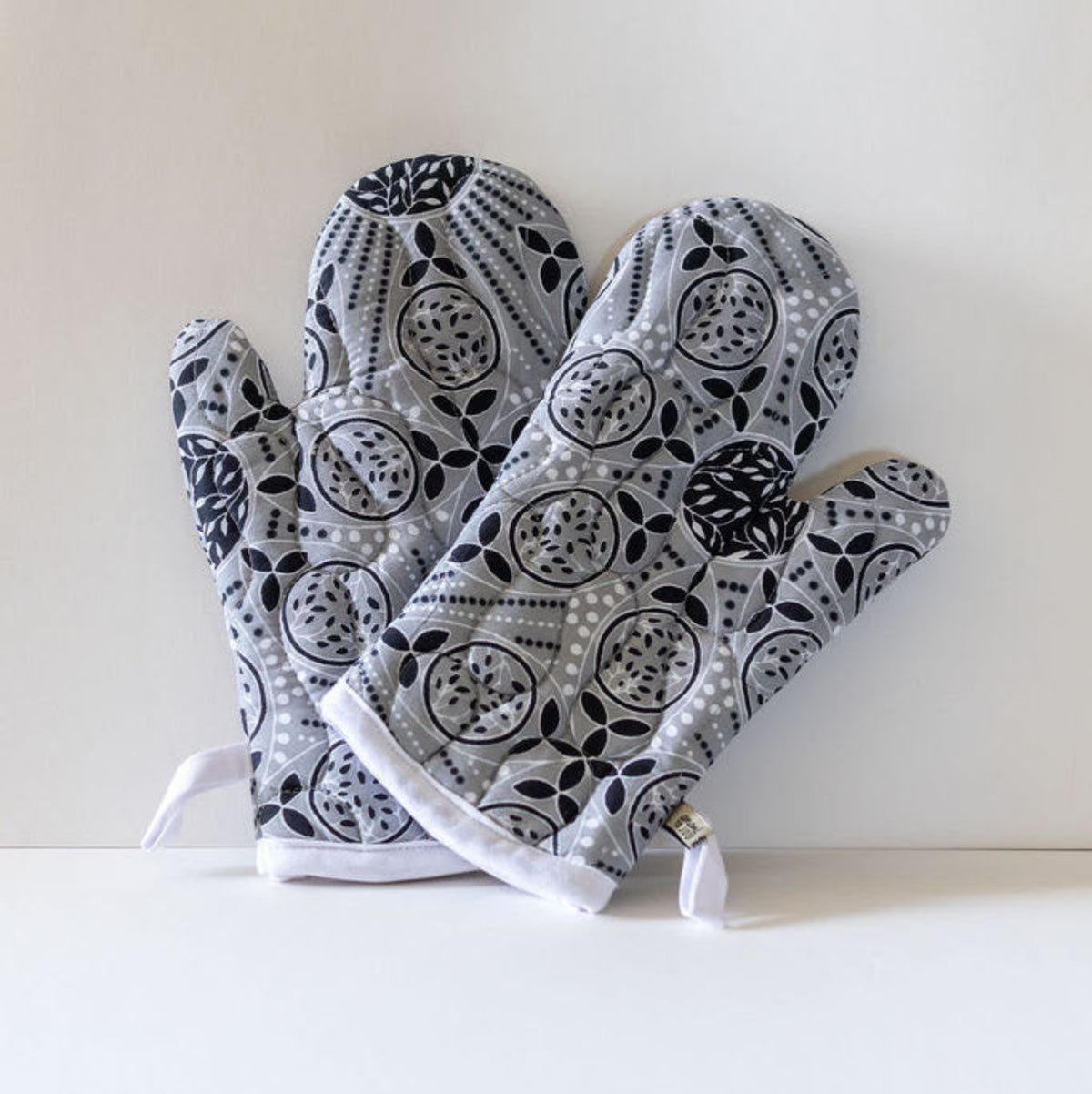 A pair of quilted oven mitts with a black, white, and gray geometric pattern, displayed on a white surface against a light beige background.