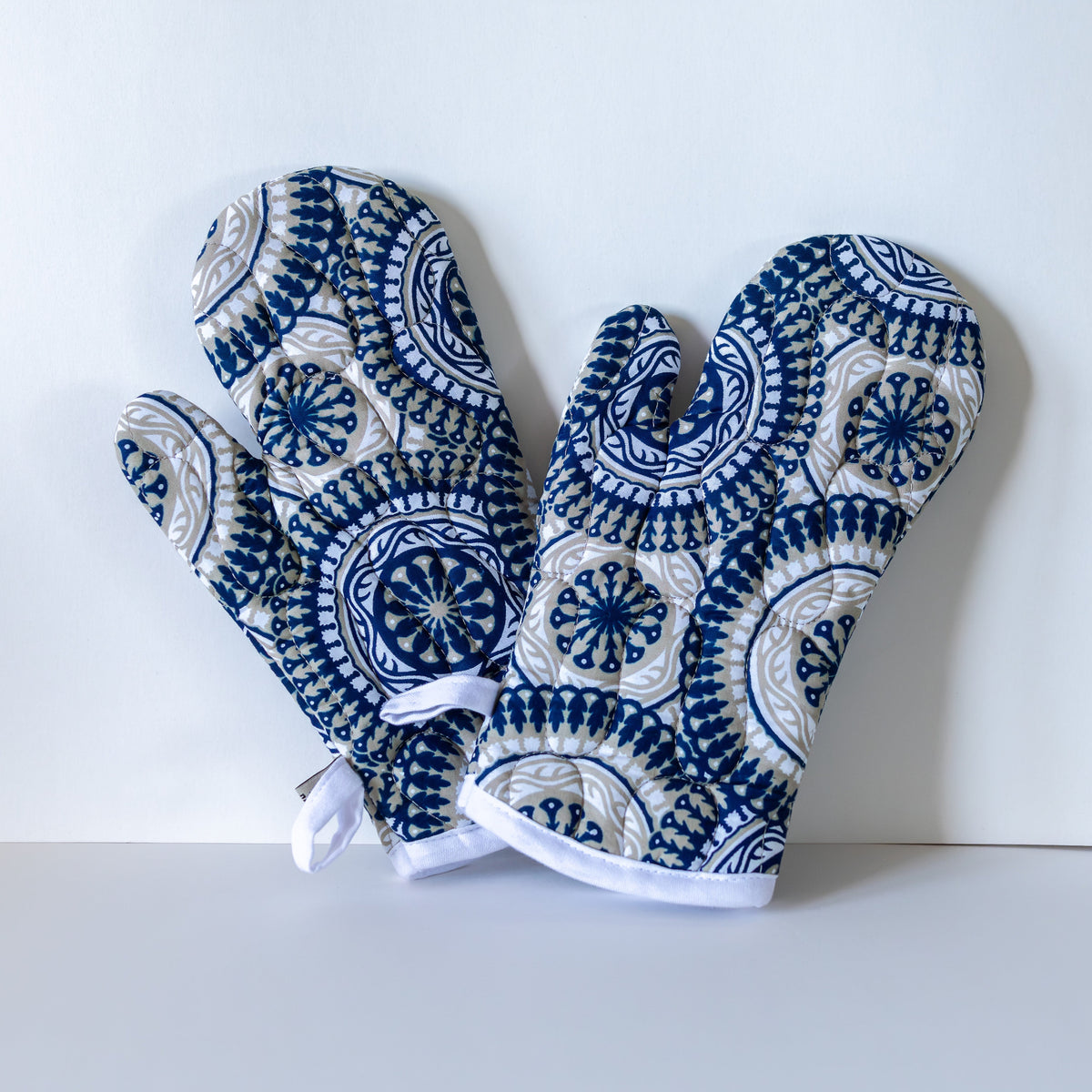 A pair of oven mitts with a navy blue, white, and beige geometric medallion pattern, displayed on a light gray surface against a white background.