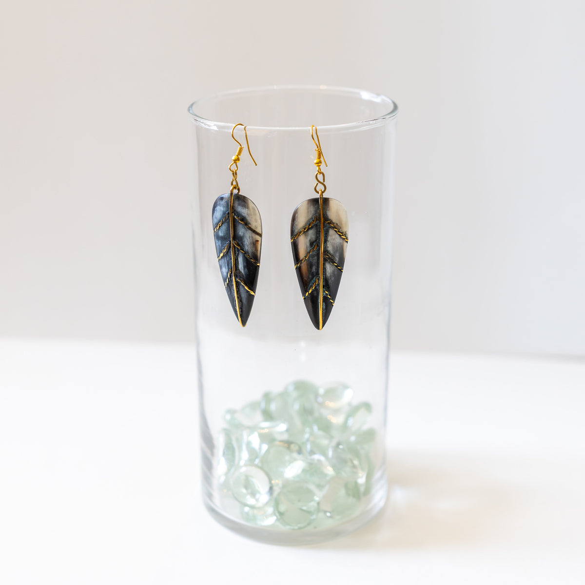 Long dark horn and brass earrings hanging from a clear glass on a white background