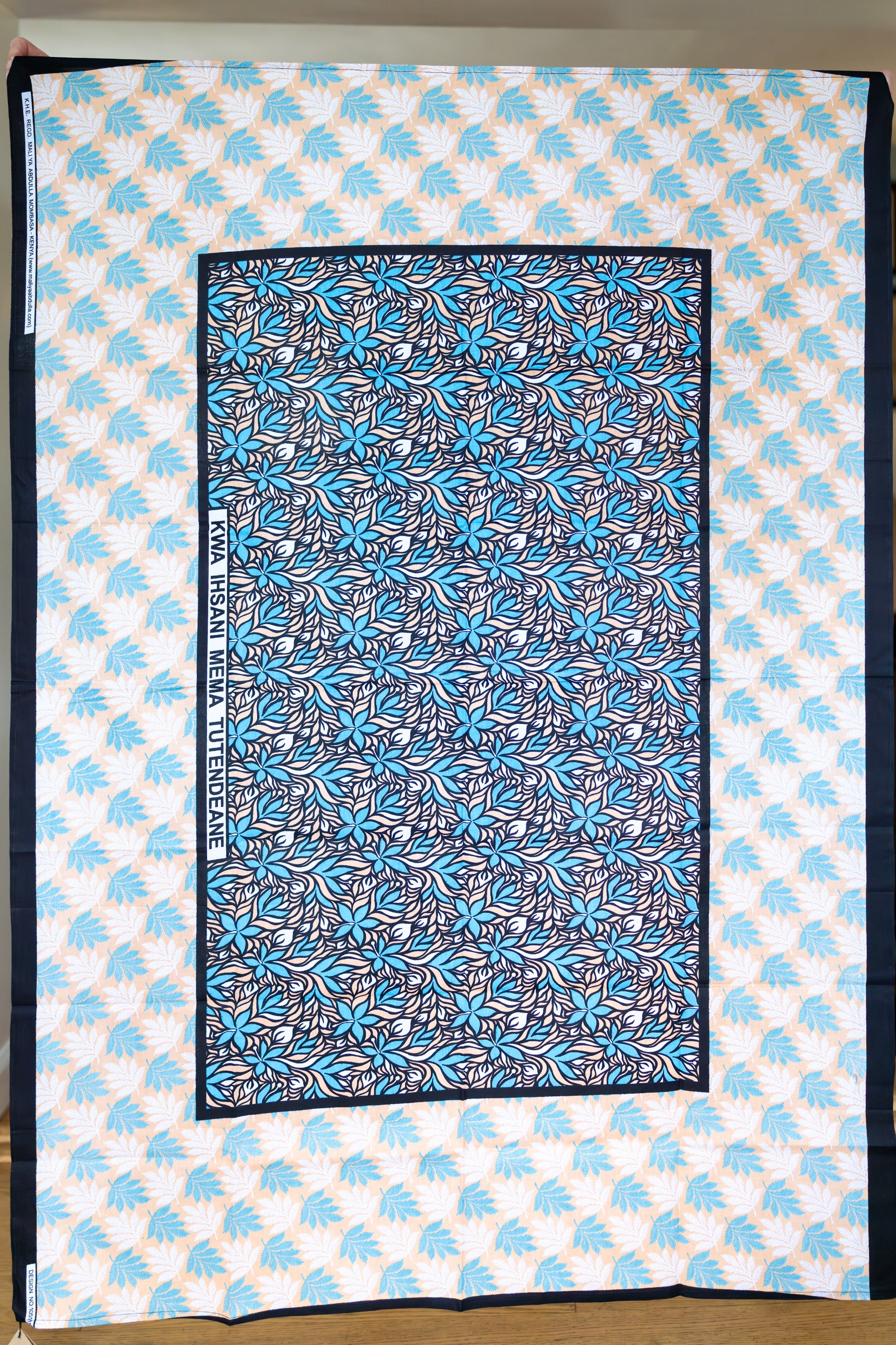 A scarf with a dense blue, black, and white floral pattern, bordered in black and displayed flat on top of a larger white fabric background with a light blue palm tree print.
