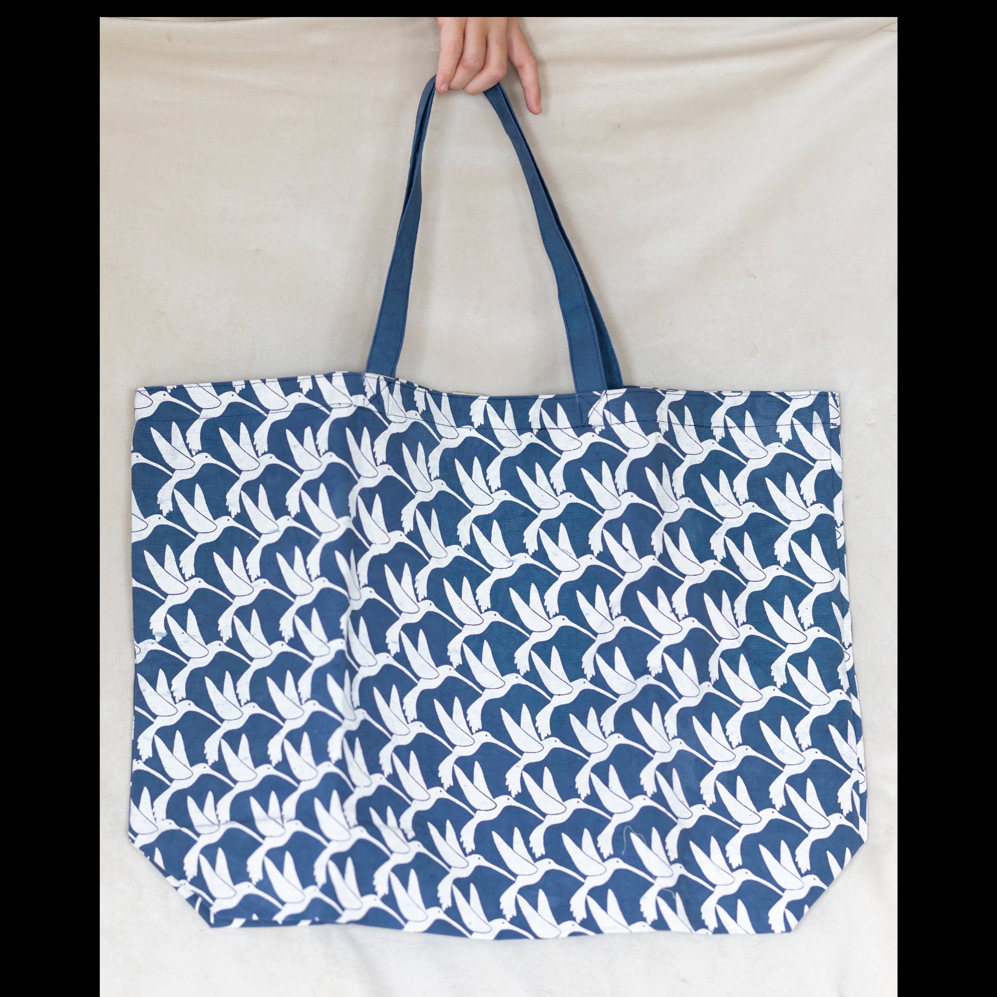 A large tote bag with a repeating white bird pattern on a navy blue background, held by a hand against a light beige fabric backdrop.