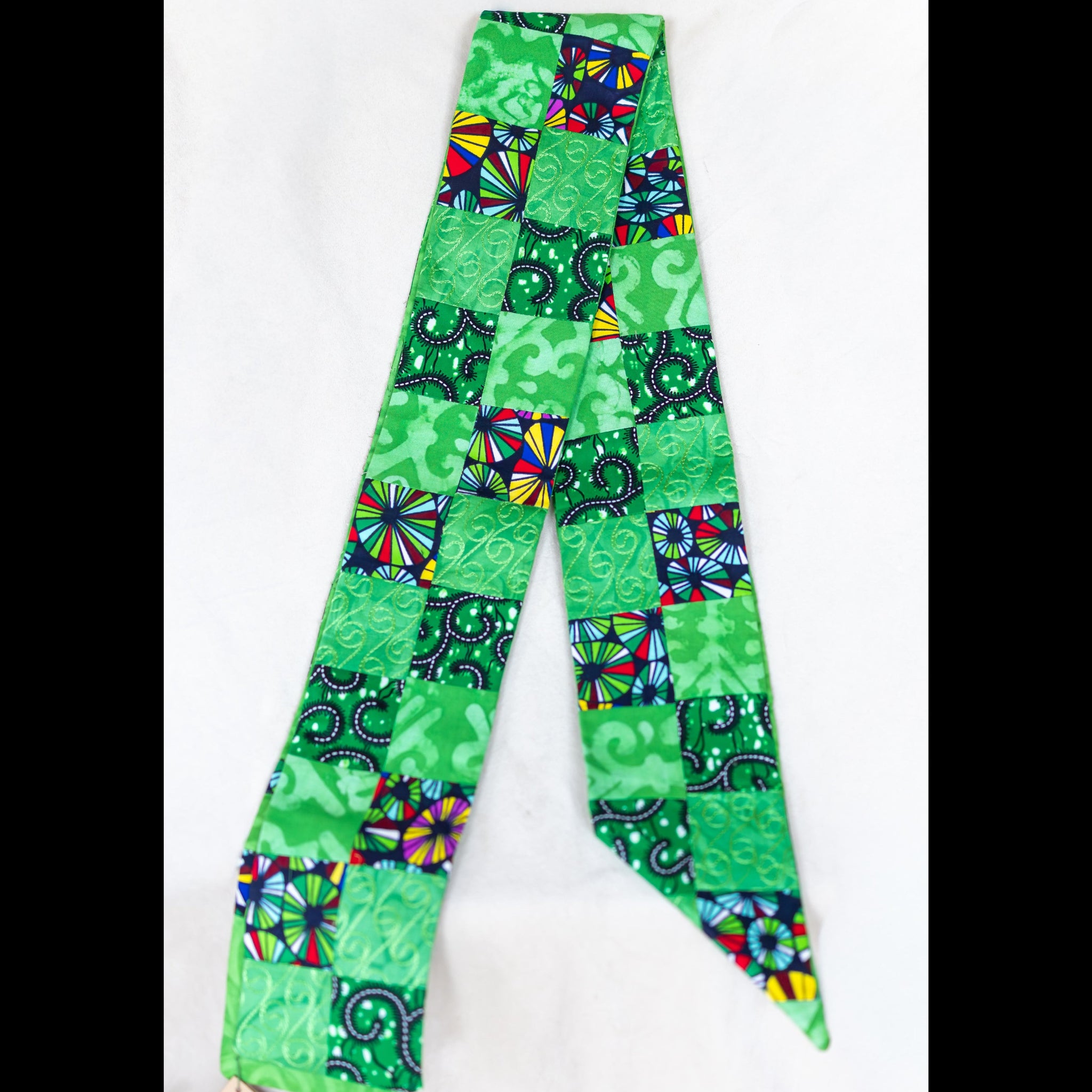 A green patterned stole with a patchwork design of various African  print squares, laid flat on a white background.