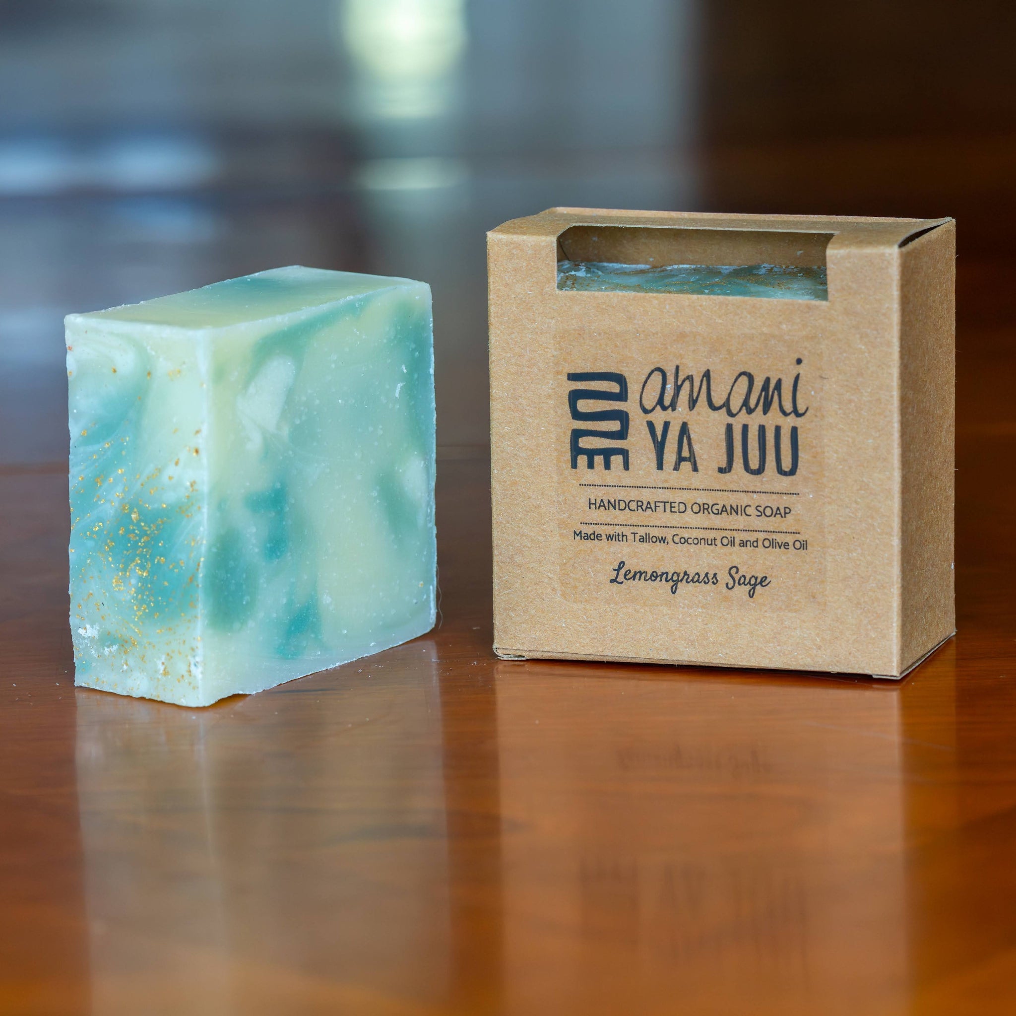 A rectangular bar of marbled light blue soap with white swirls and tiny gold flecks sits on a polished wooden table next to its kraft paper packaging box. The box is printed with the brand name 'Amani Ya Juu' and the scent 'Lemongrass Sage'.