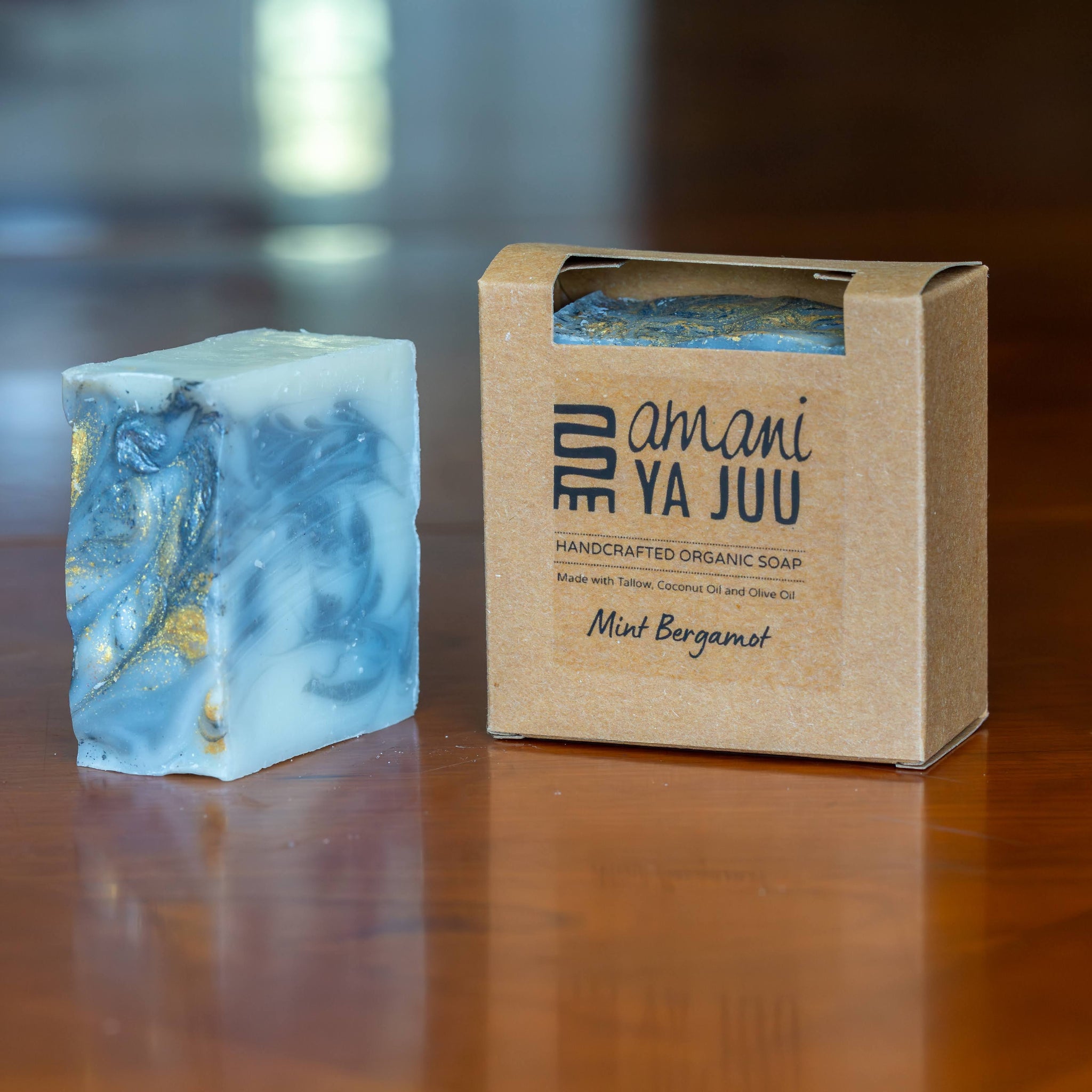 A rectangular bar of marbled blue and white soap with gold flecks stands next to its kraft paper packaging box on a polished wooden surface. The box is branded 'amani YA JUU' and labeled 'Mint Bergamot'.