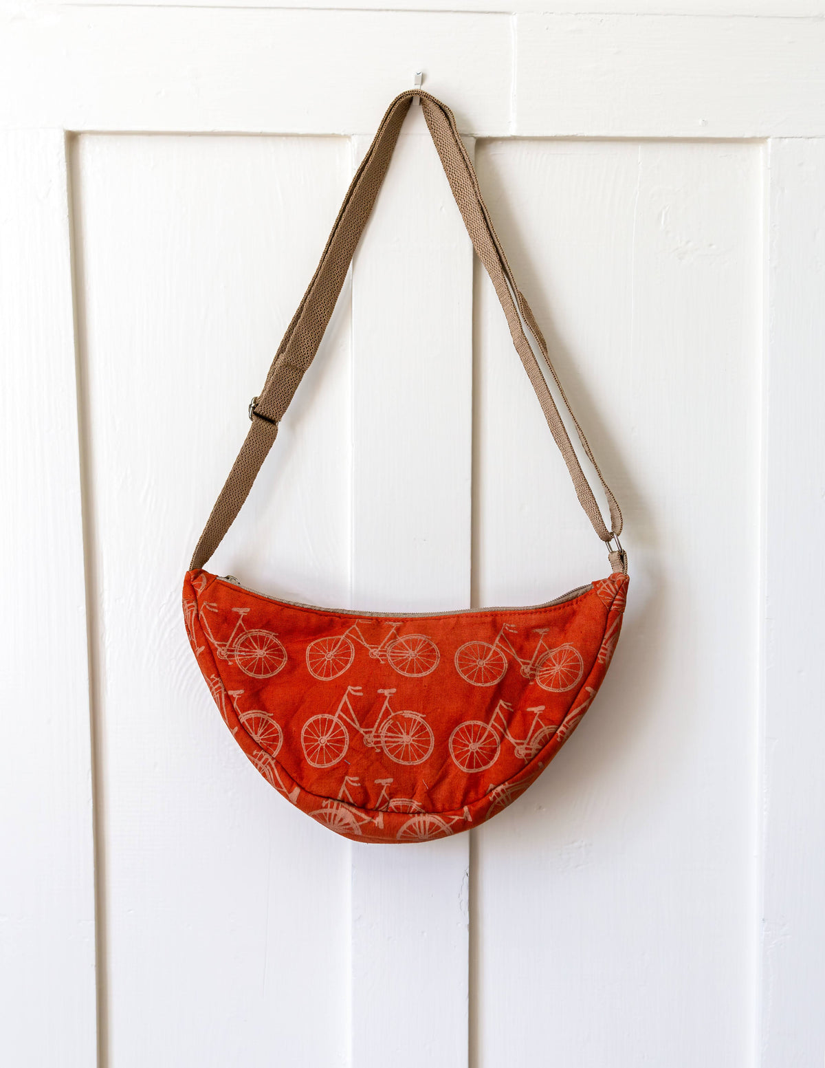 An orange crescent-shaped shoulder bag with a bicycle pattern, hanging from a tan strap on a white paneled door.