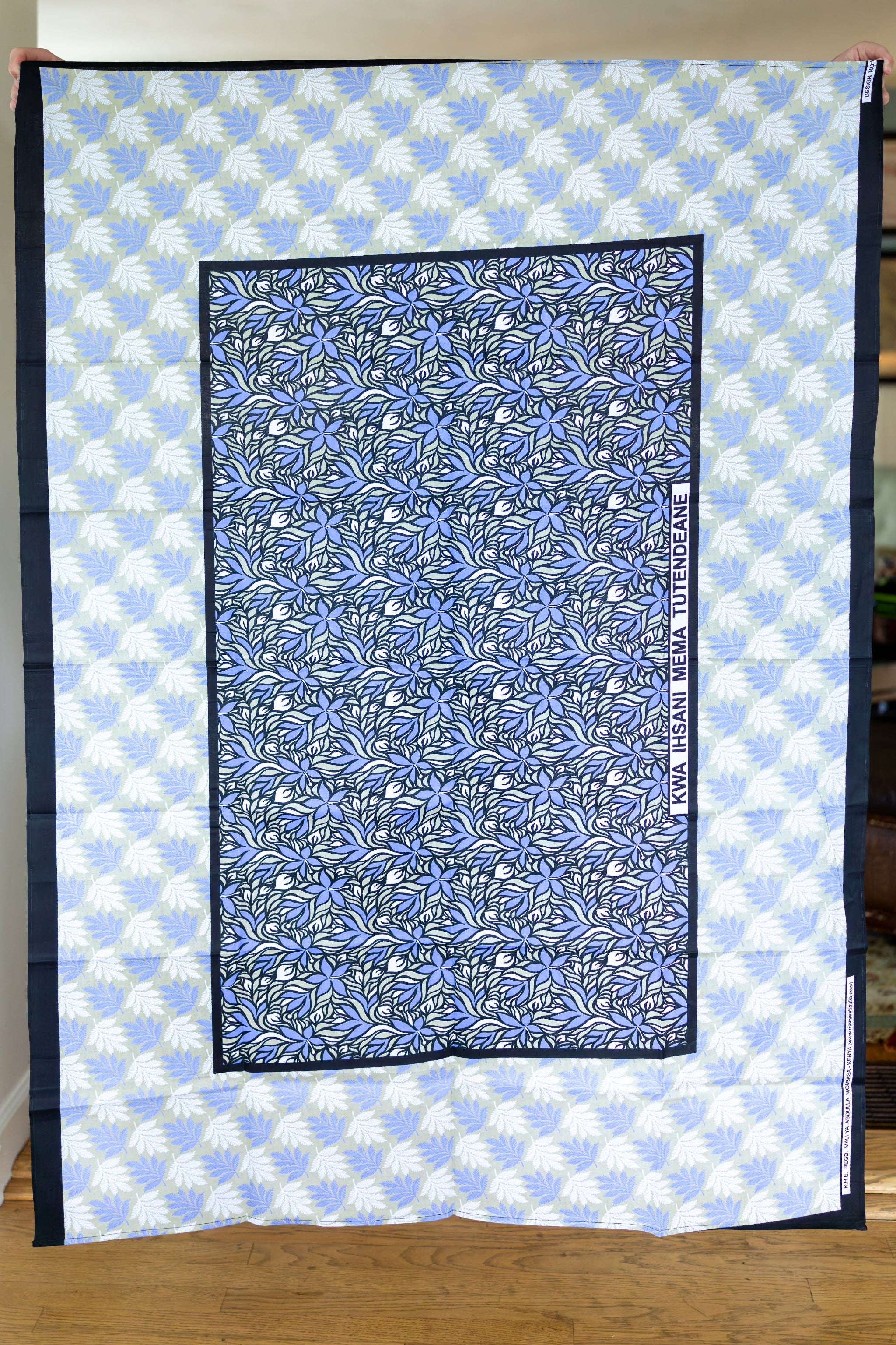 A rectangular printed textile or scarf laid flat, featuring a central periwinkle floral pattern bordered in black, surrounded by a light blue and white repeating palm-like pattern. A vertical printed text strip is visible on the right side.