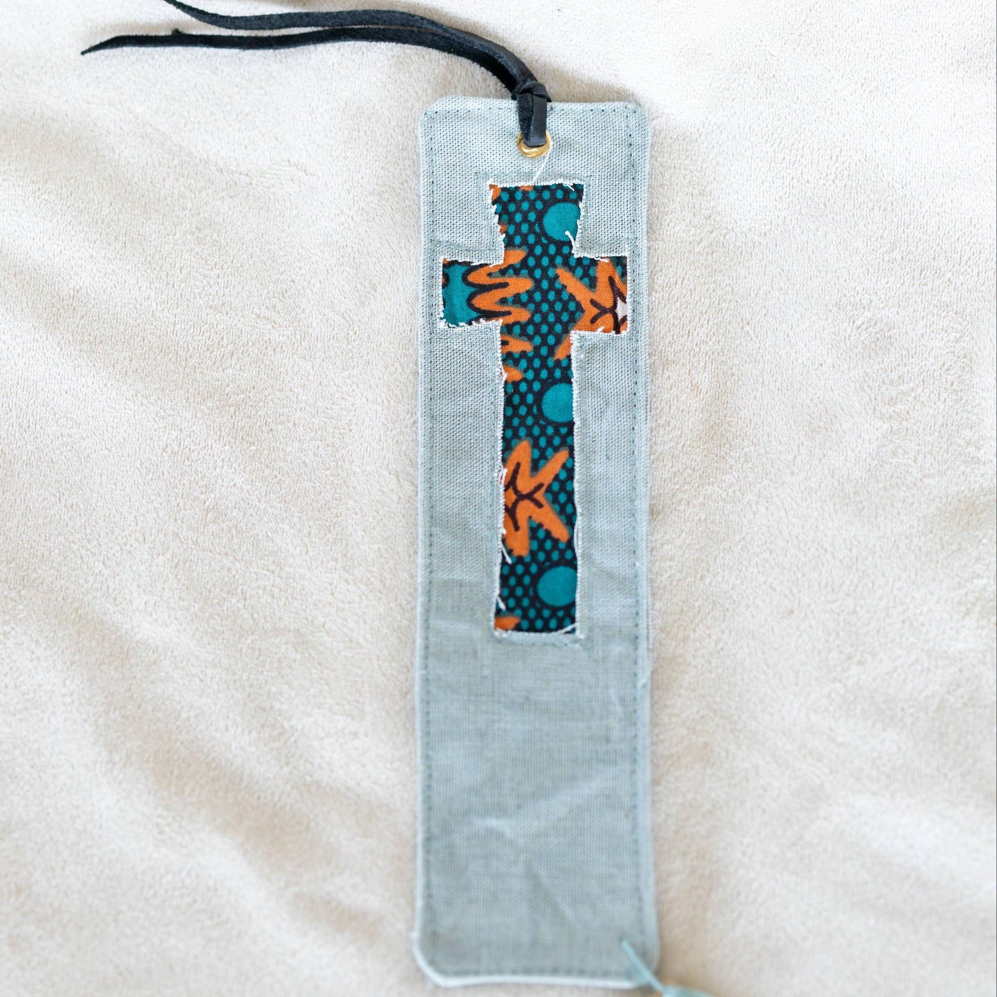 A light green fabric bookmark with a central cross-shaped patch featuring orange and teal patterned fabric, a black ribbon loop at the top, and a brown paper tag attached at the bottom, displayed on a white textured fabric background.