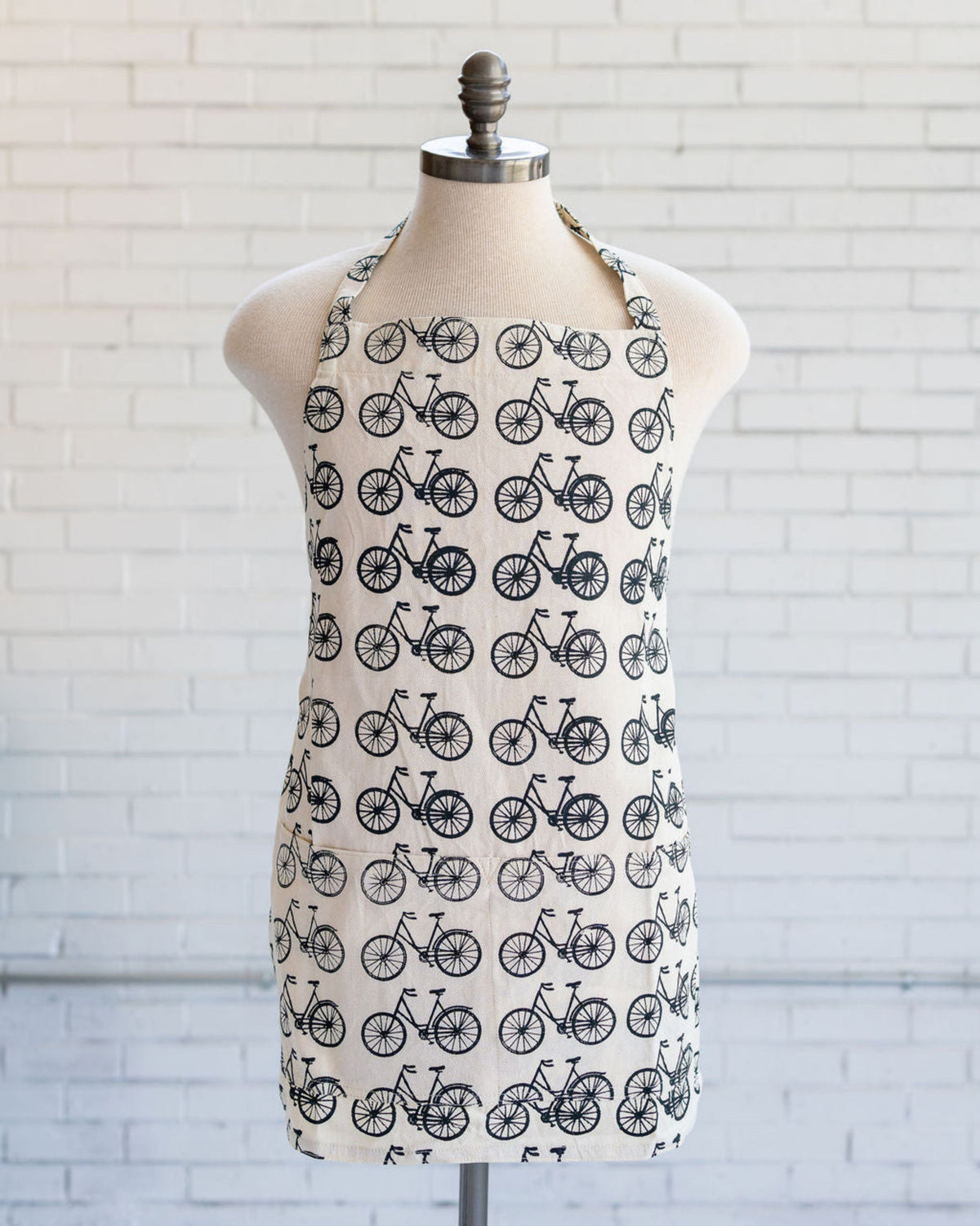A cream-colored halter apron with a black bicycle pattern, displayed on a dress form mannequin against a white brick wall background.