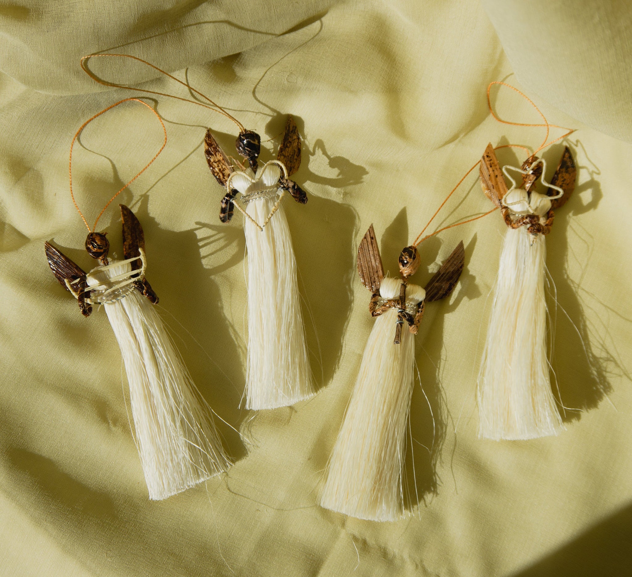 Four angel-shaped decorative items with white dresses and brown wings on a beige fabric background.