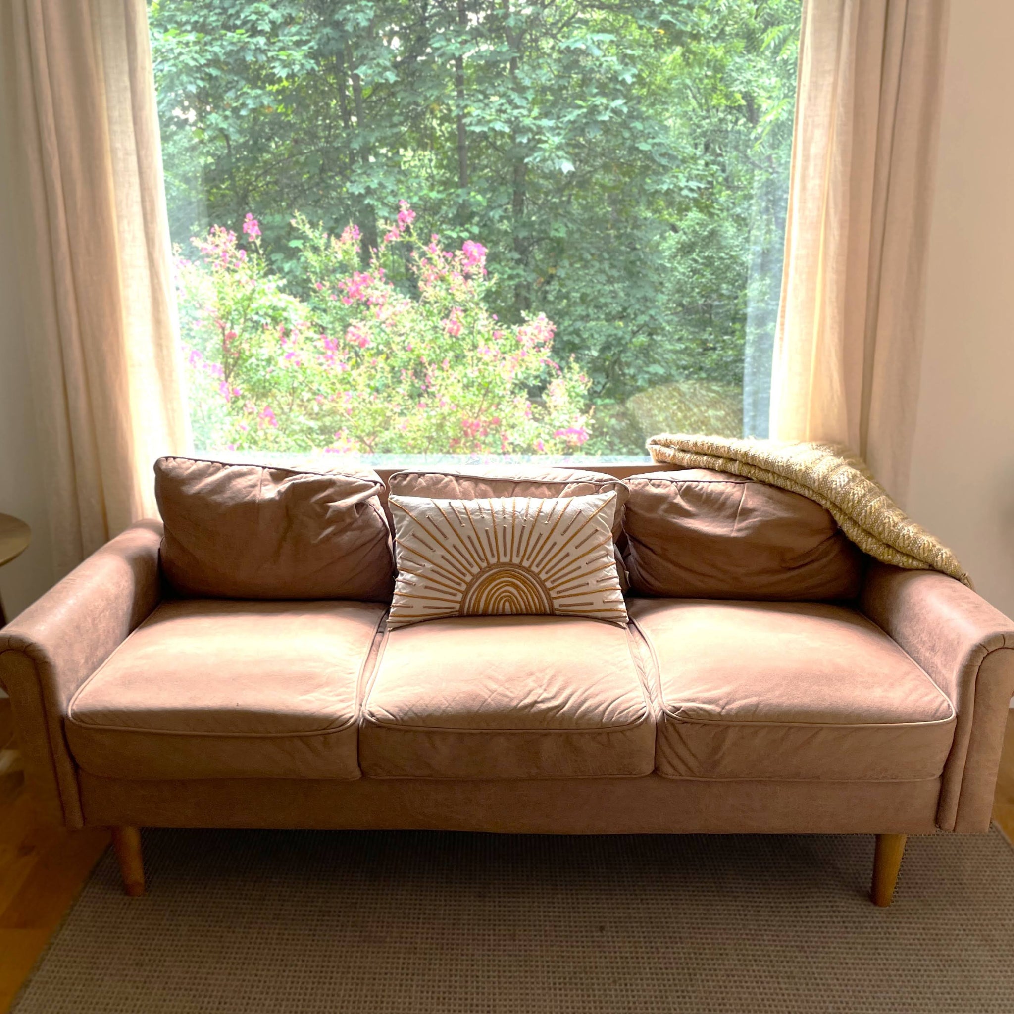 Beige sofa with a decorative pillow and blanket in front of a large window with curtains.